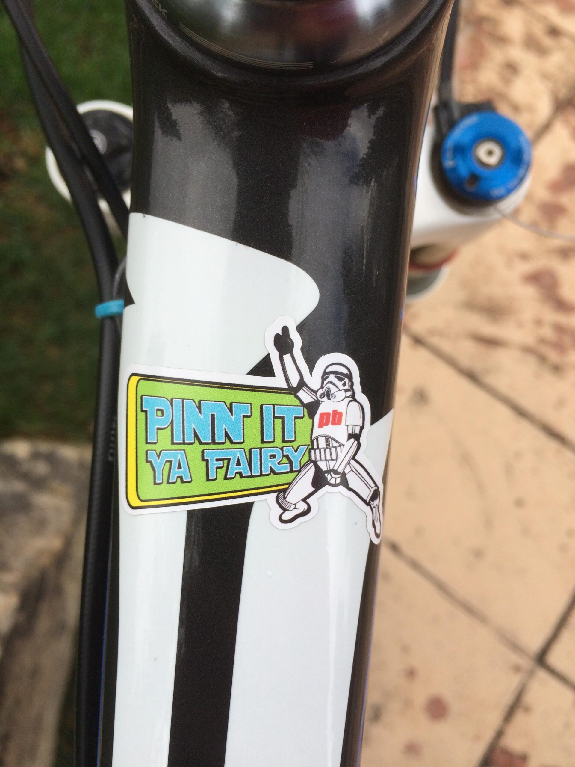 Rocky Mountain Vertex 990 rsl 2013: A close-up view of a bicycle frame featuring a colorful sticker that reads "PINN IT YA FAIRY" with an illustration of a character in a playful pose. The bike has a sleek black and white design with visible components like the handlebars and part of the ground in the background.
