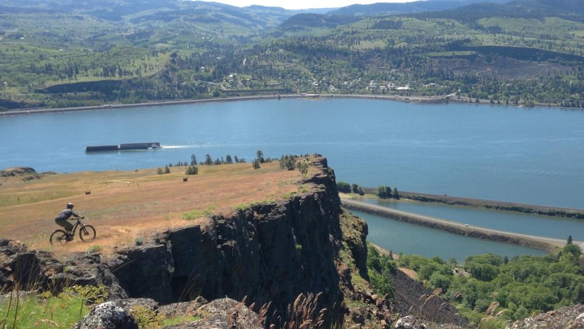 A mountain biker rides along the edge of a cliff overlooking a river surrounded by lush green hills and distant mountains on a clear day. A barge navigates the water below, while scattered trees and grassy areas add to the natural landscape. Syncline mountain bike trail.