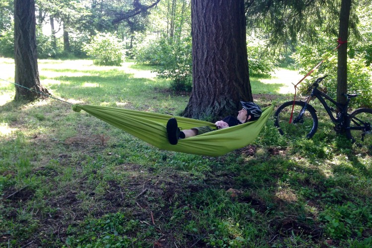 A person relaxing in a green hammock set between two trees, wearing a bicycle helmet. A mountain bike is positioned nearby, surrounded by a grassy area with trees and bushes in the background, indicating a peaceful outdoor setting.