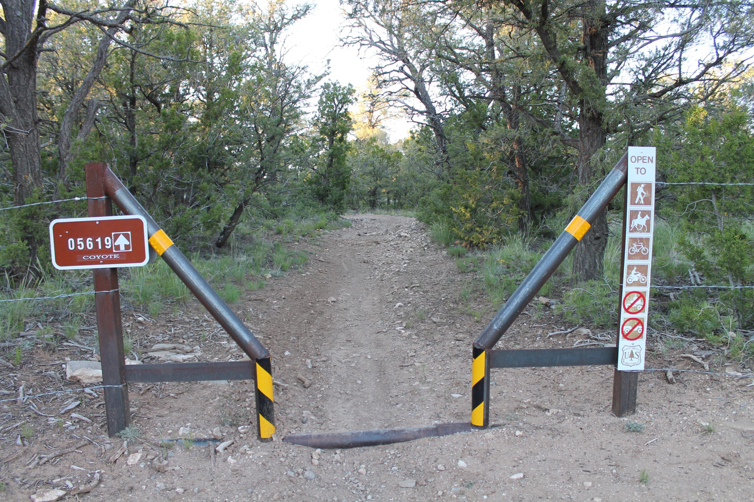 A dirt trail leading into a wooded area, marked by a gate with a brown sign labeled "05619 Coyote." Adjacent to the gate is a sign indicating the trail is open to hikers, cyclists, and horseback riders, along with various prohibited activities illustrated through symbols. The surrounding area features greenery and trees, suggesting a natural setting. Coyote Trail mountain bike trail.