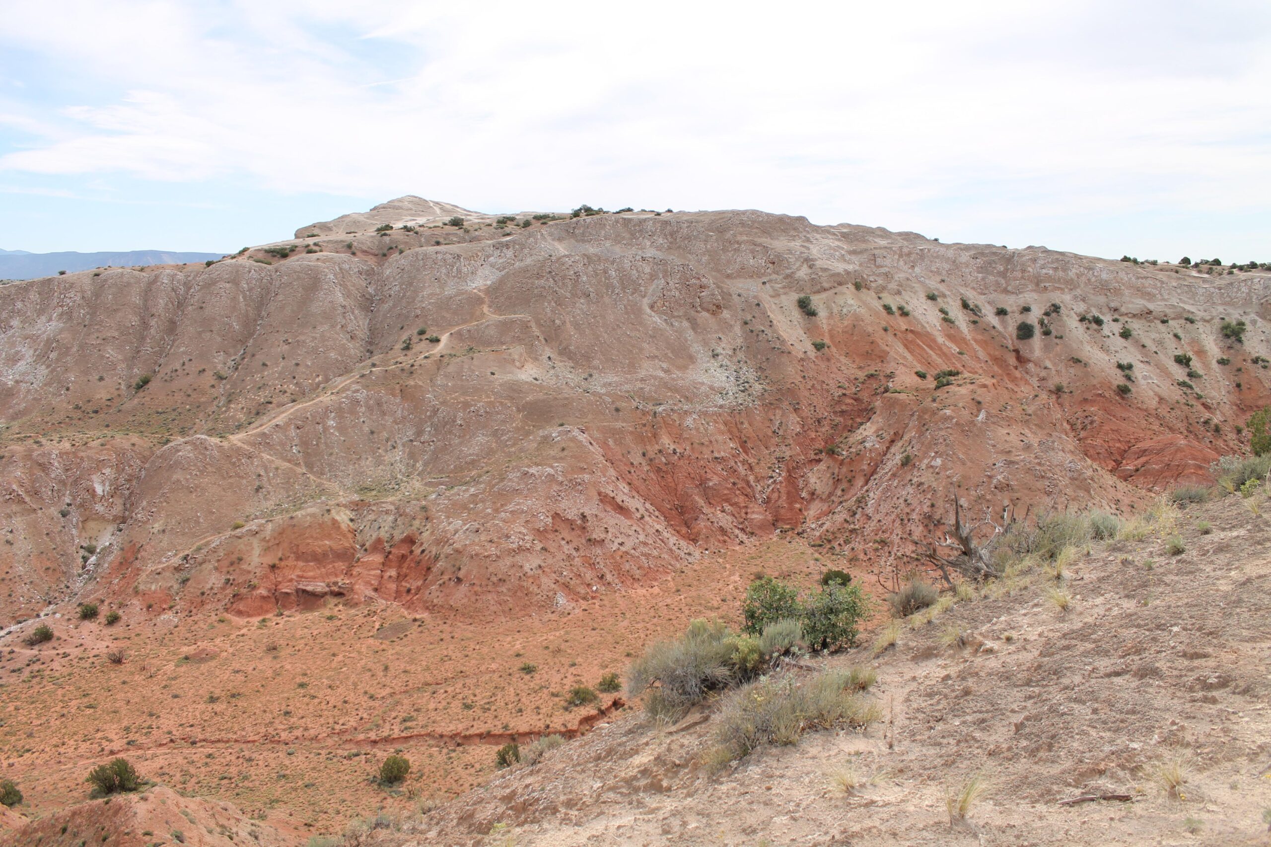 A rugged, arid landscape featuring a series of rocky, reddish-brown hills and gentle slopes, set against a cloudy sky. Sparse vegetation including small shrubs and grasses is visible in the foreground, highlighting the dry terrain. The geological layers of the hills showcase various shades and textures, indicating a rich natural history. White Ridge Bike Trails mountain bike trail.