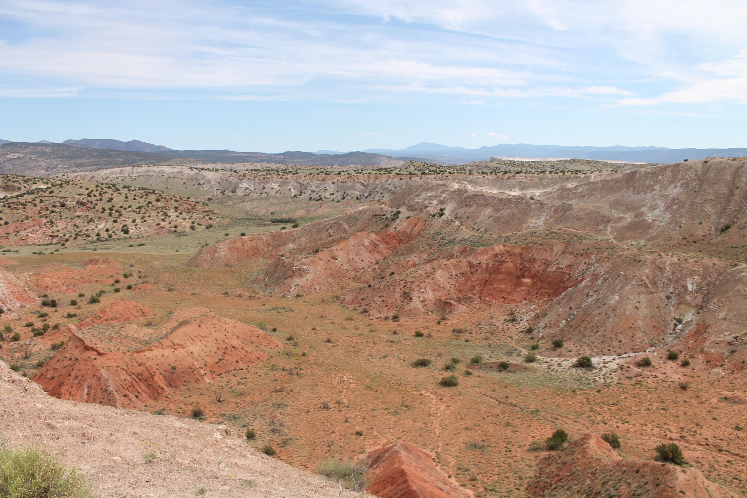 A panoramic view of a rugged landscape featuring colorful, rolling hills in shades of red, orange, and gray under a partly cloudy sky. The terrain is dotted with sparse vegetation, highlighting the dry, arid environment typical of a desert region. White Ridge Bike Trails mountain bike trail.