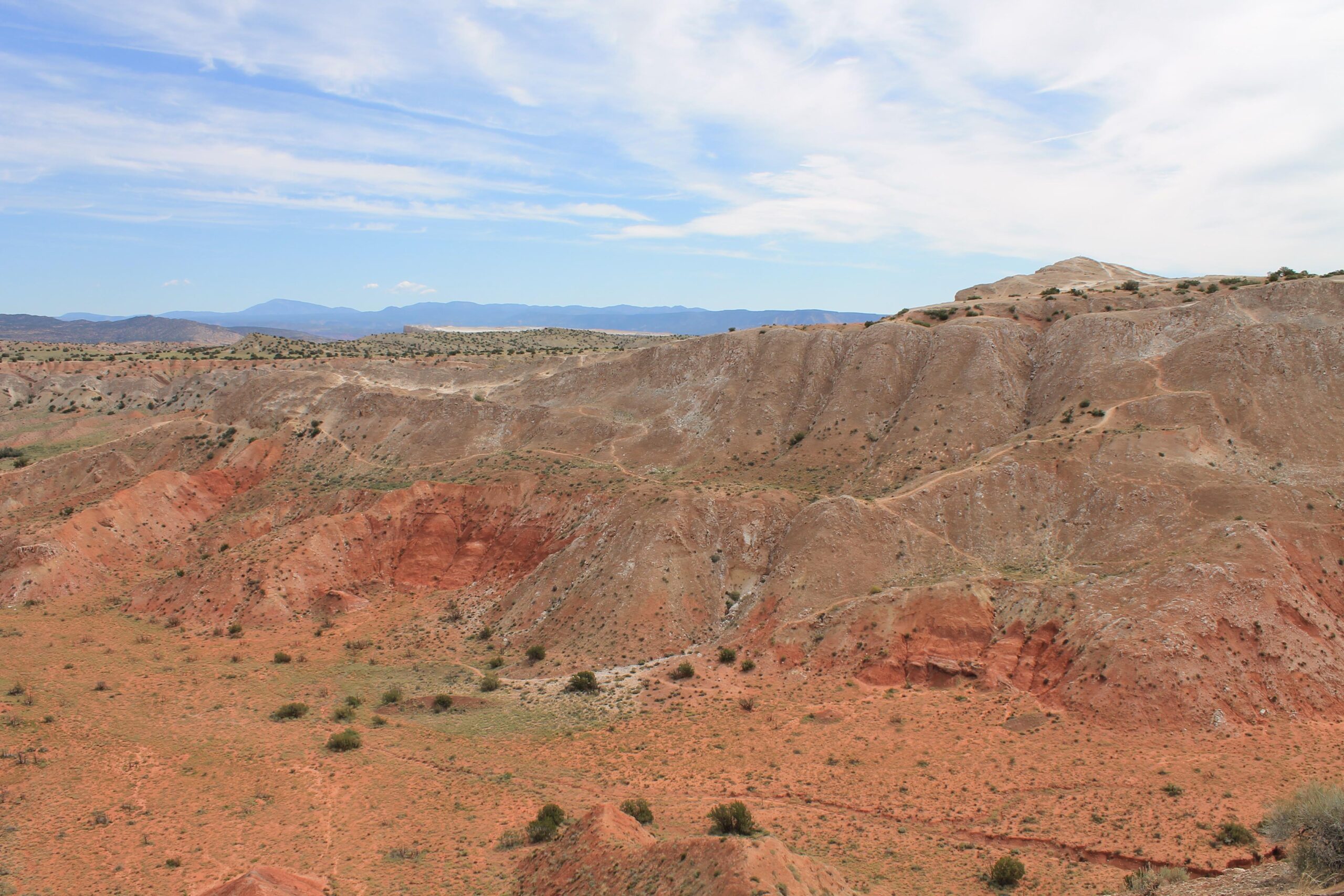 A panoramic view of rugged, colorful hills and valleys characterized by shades of red and brown soil, dotted with sparse vegetation under a clear blue sky with wispy clouds. White Ridge Bike Trails mountain bike trail.