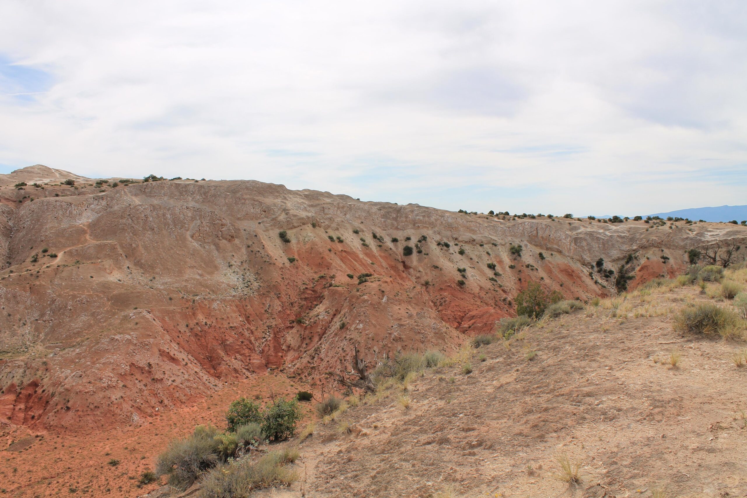 A rocky landscape featuring layers of reddish-brown earth and sparse vegetation, with a cloudy sky overhead. The scene captures the undulating topography of a canyon or hillside, showcasing the natural erosion patterns of the terrain. White Ridge Bike Trails mountain bike trail.