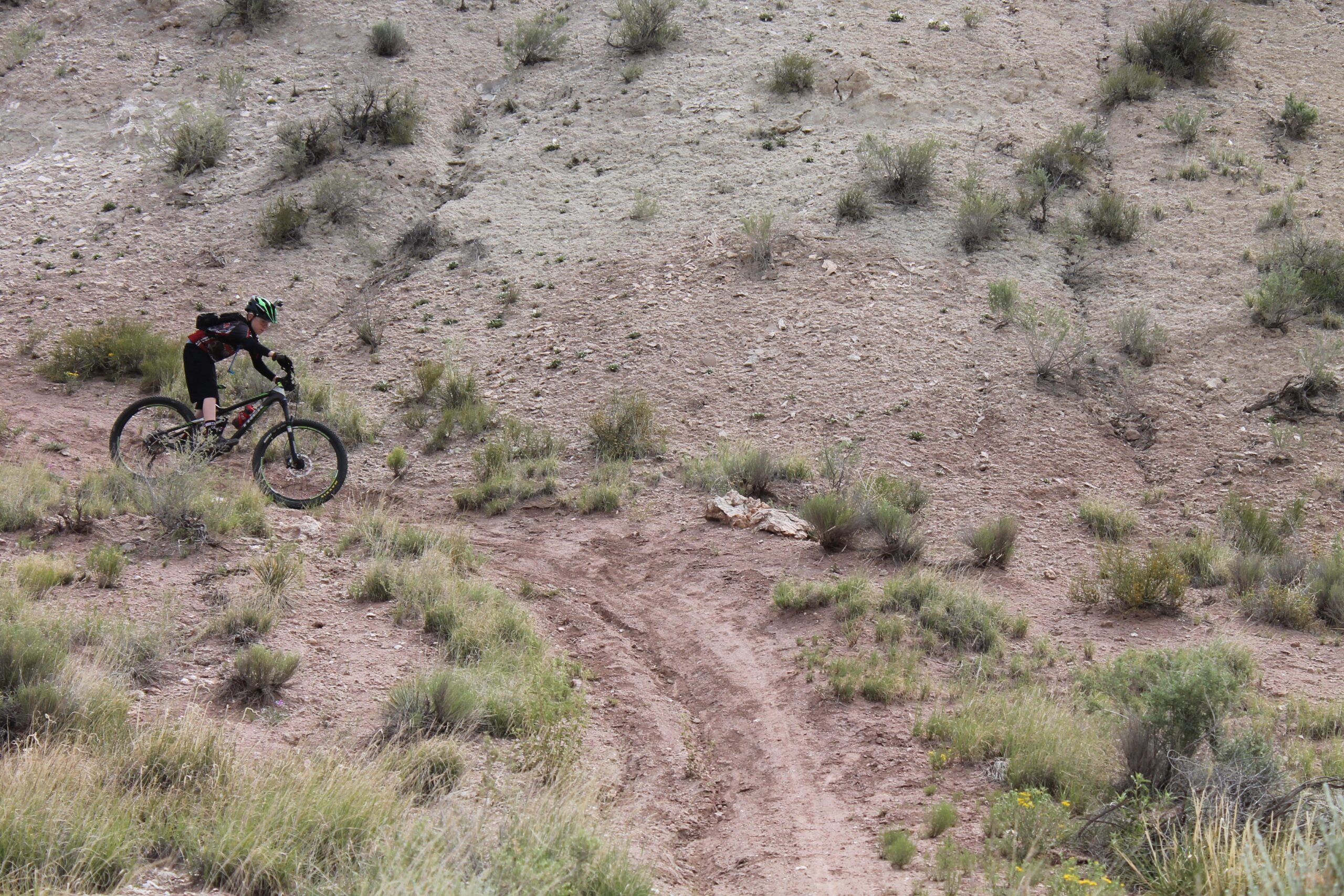 A mountain biker riding along a dirt trail on a hillside, surrounded by sparse vegetation and rocky terrain. The rider is dressed in a black outfit and a helmet, navigating the winding path through a natural landscape. White Ridge Bike Trails mountain bike trail.