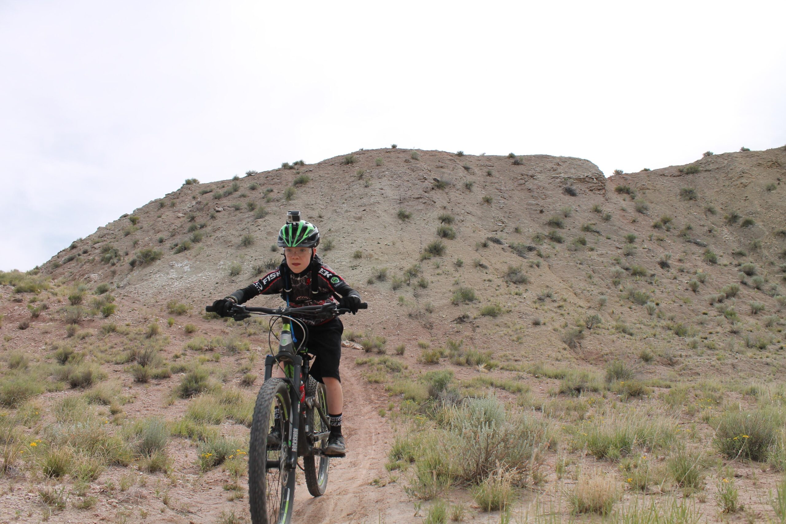 A young cyclist riding a mountain bike on a dirt trail in a hilly, arid landscape. The rider is wearing a black and green helmet and gear, with a camera mounted on the helmet, focused on navigating the trail. The background features a sloped hill with sparse vegetation and a cloudy sky. White Ridge Bike Trails mountain bike trail.