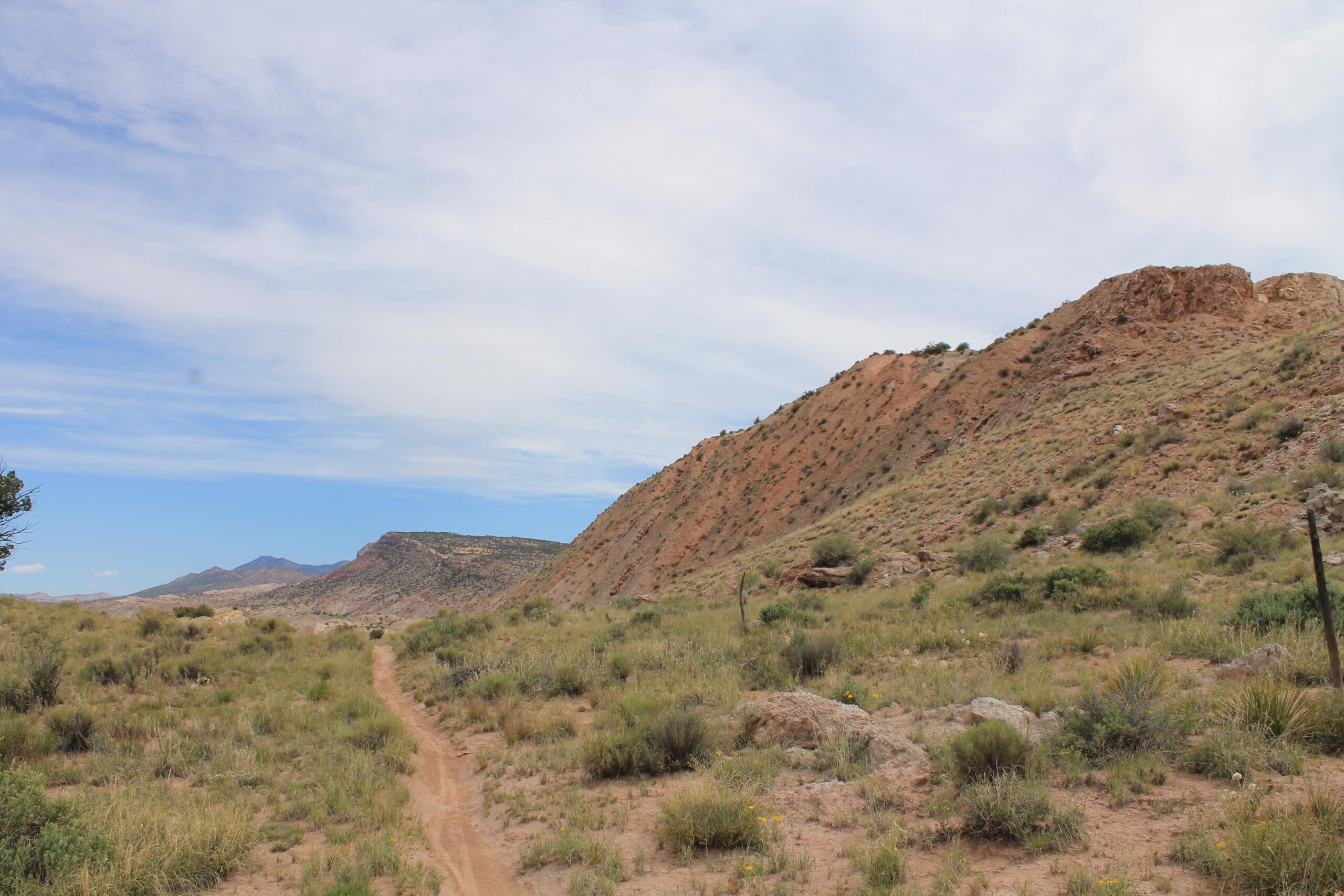 A dirt path winds through a landscape of dry grasses and low shrubs, leading towards a rocky hillside under a partly cloudy sky. The terrain features rolling hills and distant mountains in the background. White Ridge Bike Trails mountain bike trail.