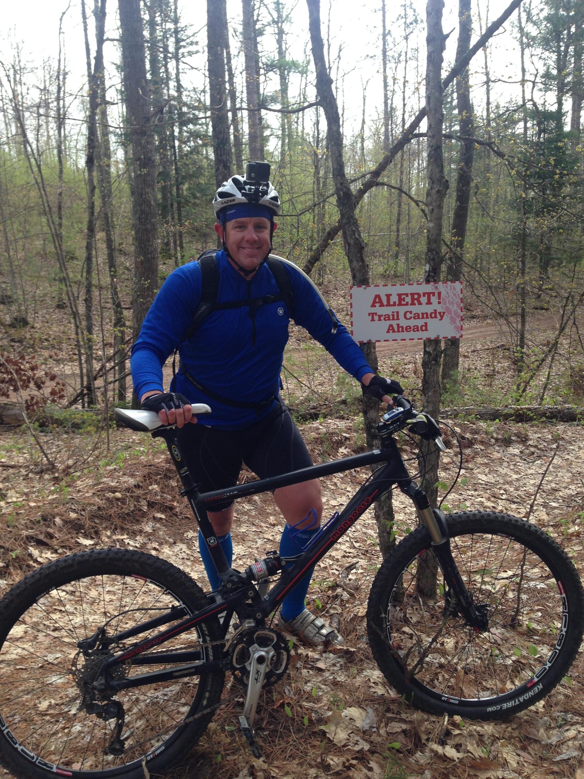A person wearing a blue long-sleeve shirt, cycling shorts, and a helmet with a camera mounted on it stands beside a mountain bike on a wooded trail. In the background, a sign reads, "ALERT! Trail Candy Ahead." The surrounding environment features trees and leaf litter, indicating a natural outdoor setting. Seeley Pass Trail mountain bike trail.
