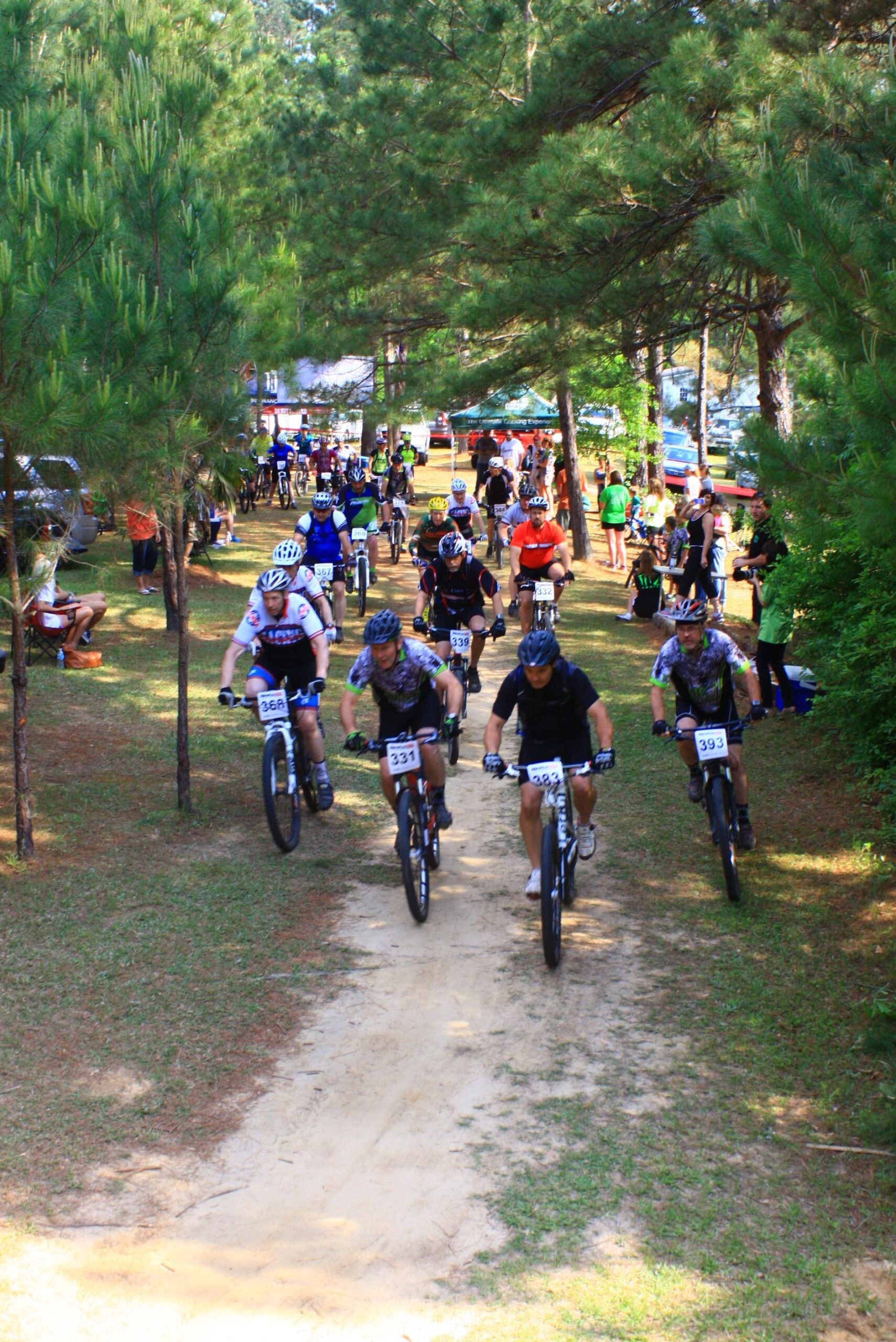 A group of mountain bikers riding on a dirt path through a wooded area during a biking event. The scene includes both competitors wearing helmets and jerseys, as well as spectators in the background. Tall pine trees line the path, and various colorful tents are visible in the distance. Mt. Zion Bike Trails mountain bike trail.