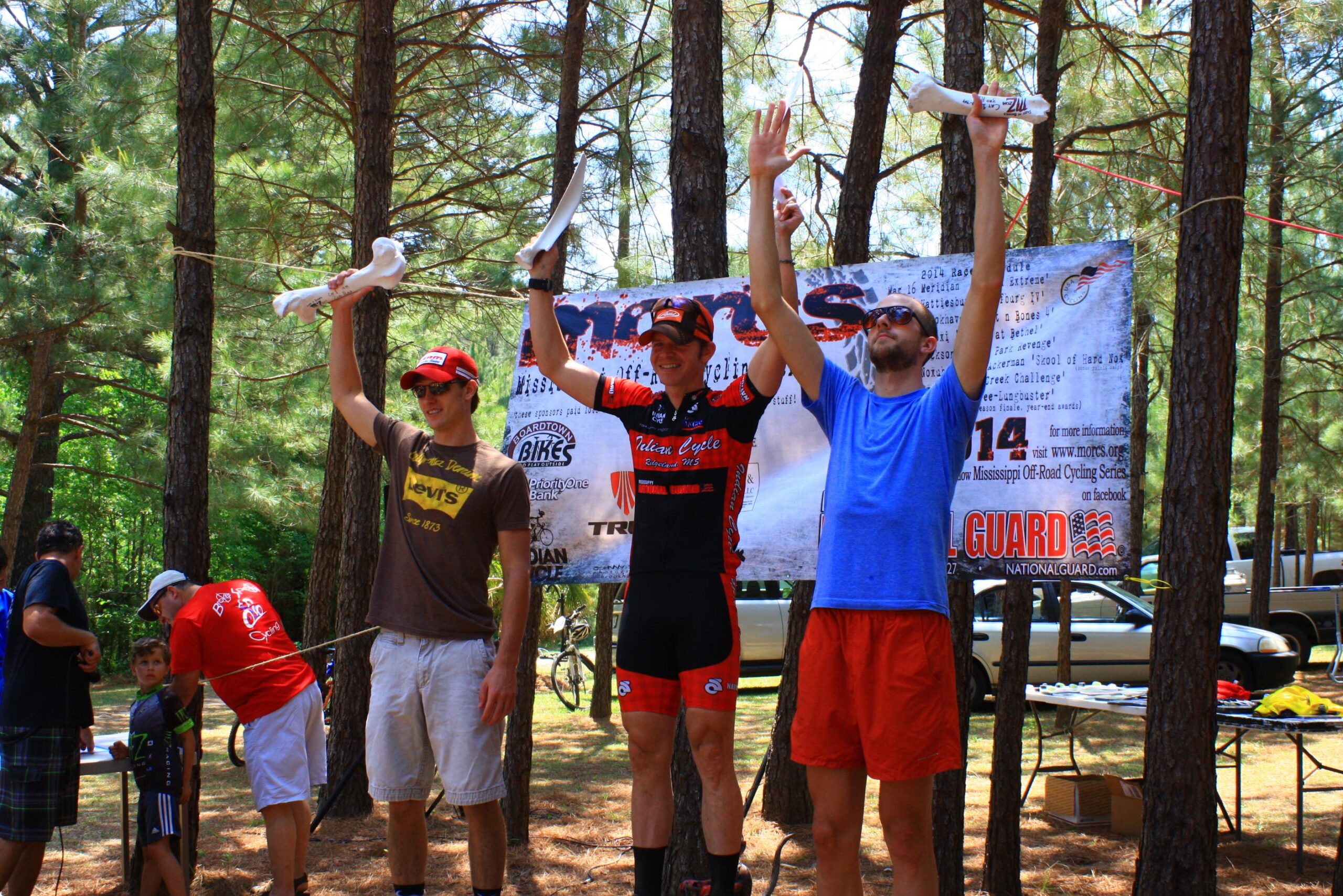 Three participants stand on a podium in a forested area, celebrating an event. The central figure, dressed in a red and black cycling jersey, holds a trophy high above their head, while the others, wearing casual clothing, also raise their hands in celebration. In the background, a banner with event details is displayed, along with spectators and bicycles nearby. Mt. Zion Bike Trails mountain bike trail.