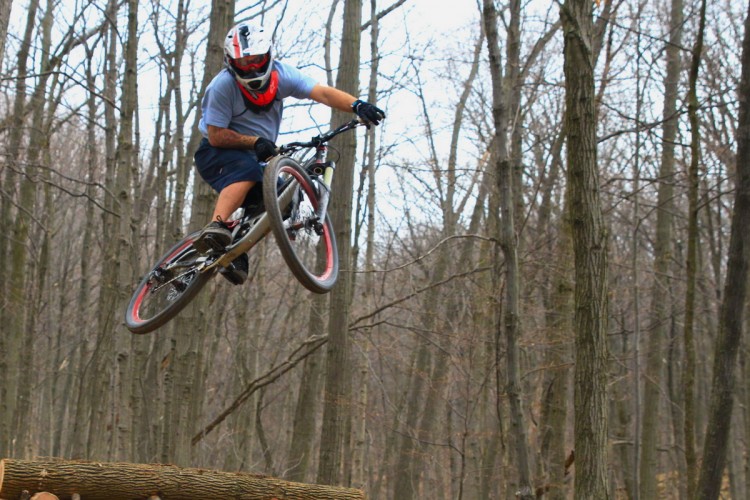 A mountain biker performing a jump over a wooden ramp in a forested area, surrounded by bare trees. The rider is wearing a helmet, goggles, and protective gear, showcasing an action-packed moment in a natural setting.