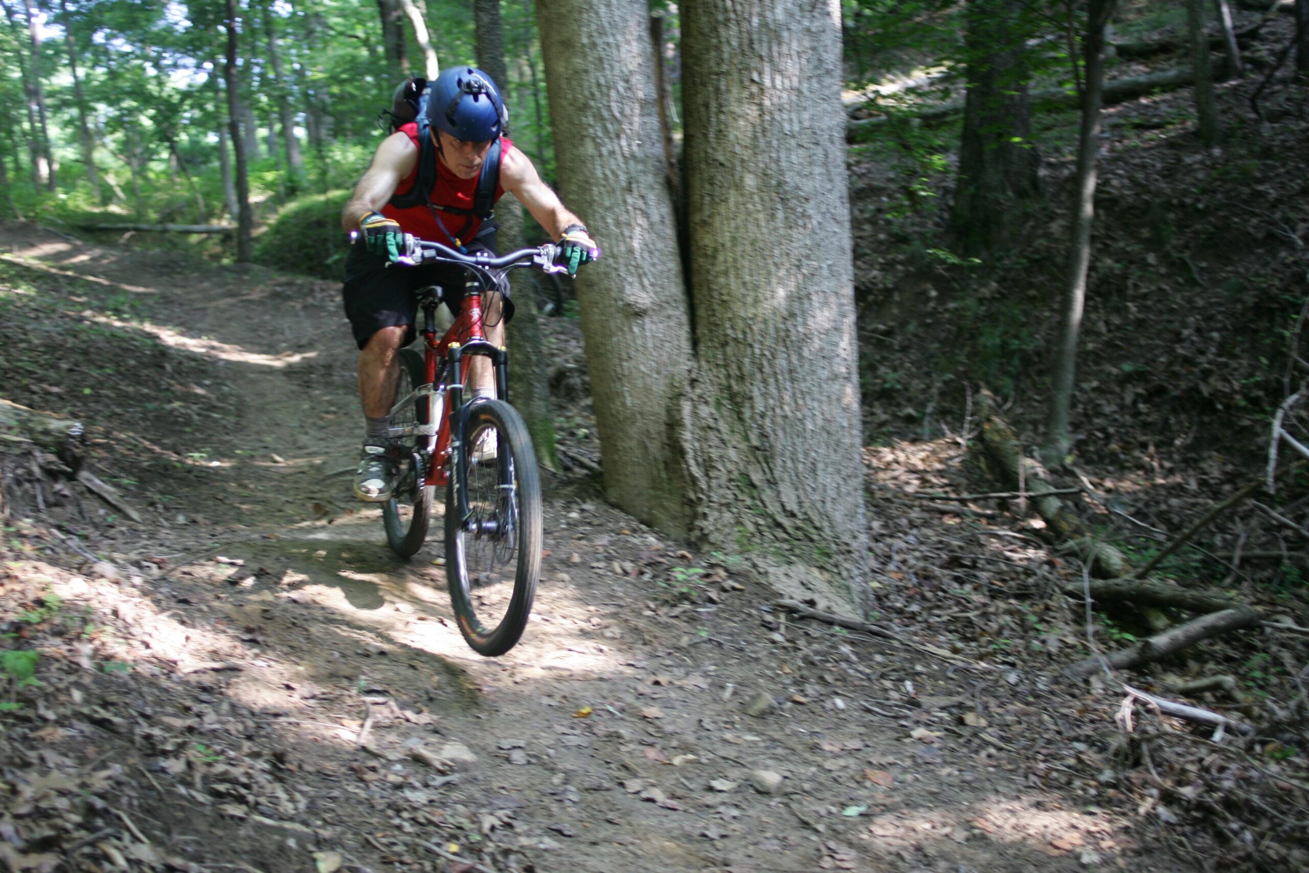 A mountain biker dressed in a red tank top and black shorts rides along a narrow dirt trail in a wooded area. He is focused on navigating the path, which is surrounded by trees and scattered leaves. Bright sunlight filters through the foliage, creating a dappled effect on the ground. Angler's Ridge mountain bike trail.