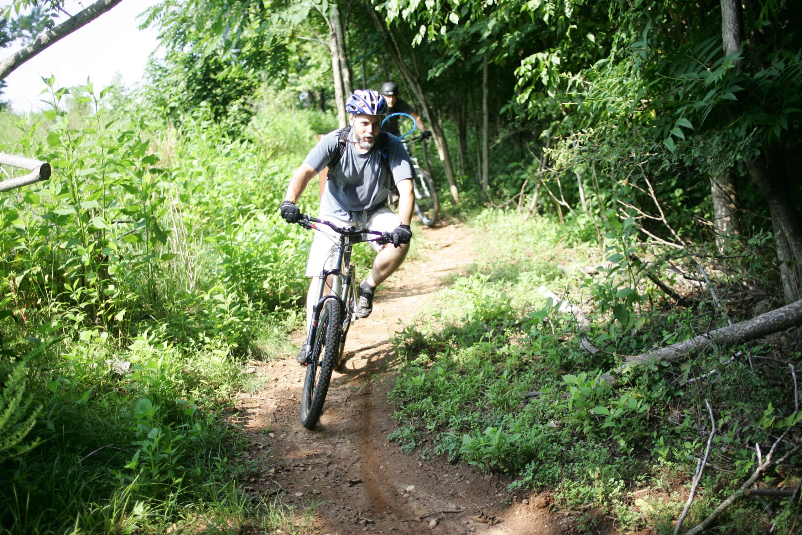 A person mountain biking on a dirt trail surrounded by lush greenery and trees, wearing a helmet and gloves, with another cyclist visible in the background. Angler's Ridge mountain bike trail.