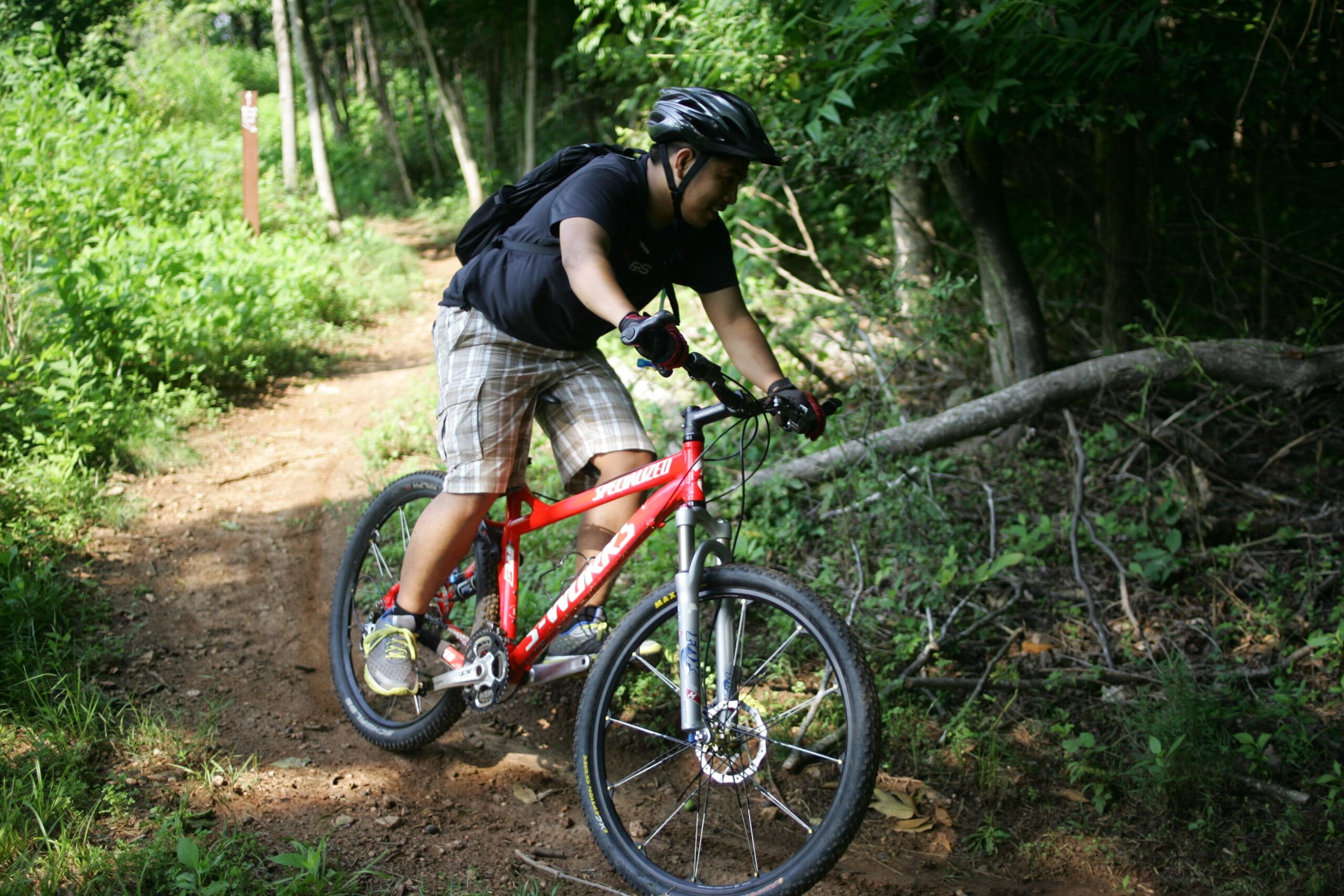 A person riding a red mountain bike on a dirt trail surrounded by lush greenery and trees, wearing a helmet and gloves for safety. Angler's Ridge mountain bike trail.