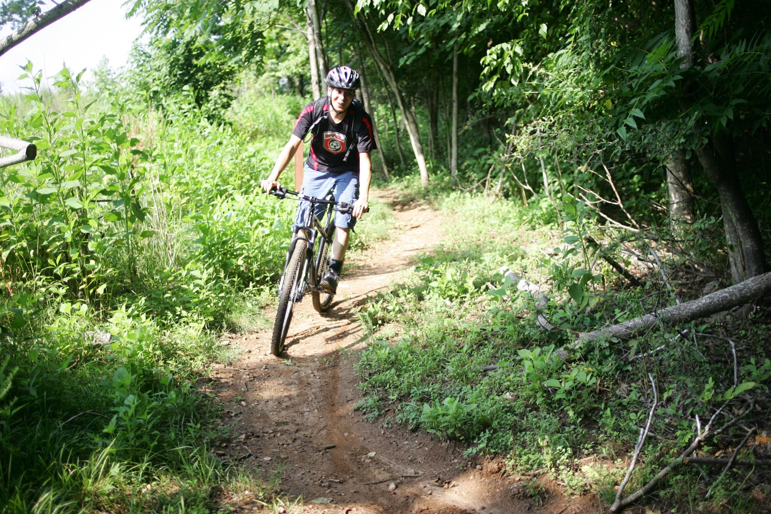 A person riding a mountain bike along a dirt trail surrounded by lush green vegetation and trees on a sunny day. The rider is wearing a helmet and casual athletic clothing, appearing focused on navigating the path. Angler's Ridge mountain bike trail.