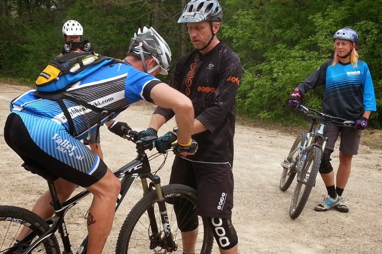 A group of three mountain bikers is gathered in a wooded area. One cyclist, wearing a blue jersey and black shorts, is leaning over his bike while another cyclist in a black shirt offers assistance. A third cyclist stands nearby, observing, and is dressed in casual biking gear. The scene captures a moment of bike maintenance or adjustment in an outdoor setting.
