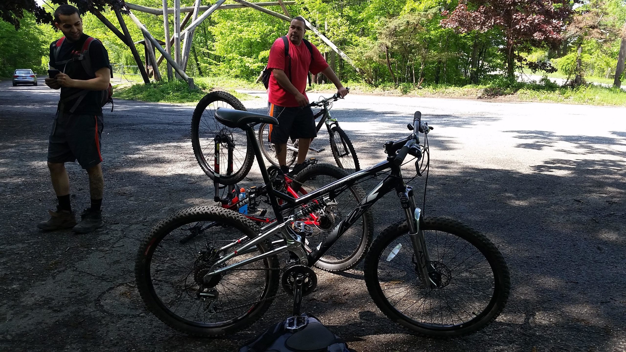Two men are near a gravel path, each standing beside a mountain bike. One man is wearing a black shirt and shorts, looking at a device, while the other is in a red shirt, facing the camera. The background features green trees and a clear road. Long Pond Ironworks State Park mountain bike trail.