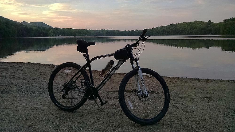 A black mountain bike stands on the sandy shore of a calm lake, with a backdrop of lush green trees and hills under a soft, pastel-hued sky at sunset. Moreau Lake State Park mountain bike trail.