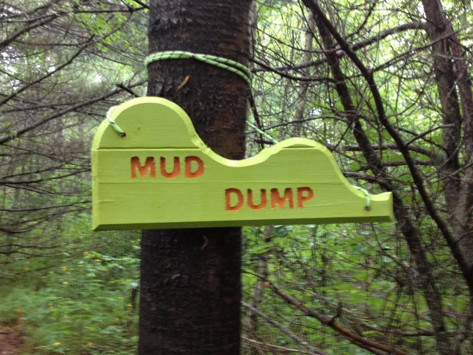 A brightly colored green wooden sign that reads "MUD DUMP," hanging on a tree in a dense forest setting. The background is filled with green foliage and various trees, creating a natural and rustic atmosphere. Mud Dump mountain bike trail.