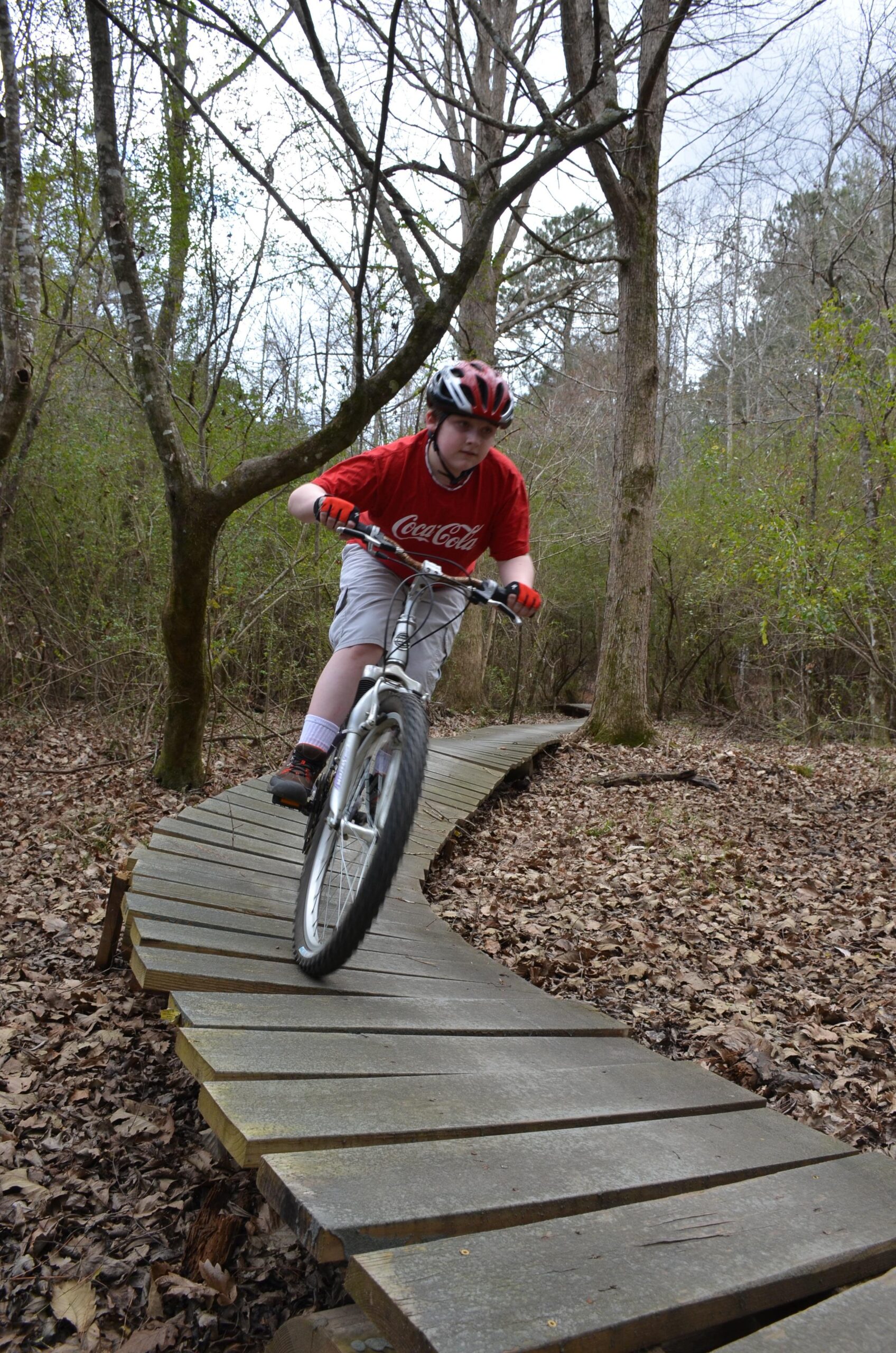 A young person riding a mountain bike on a wooden pathway through a wooded area, leaning to the side for balance. The rider is dressed in a red shirt and a helmet, with surrounding trees and fallen leaves visible in the background. Mt. Zion Bike Trails mountain bike trail.