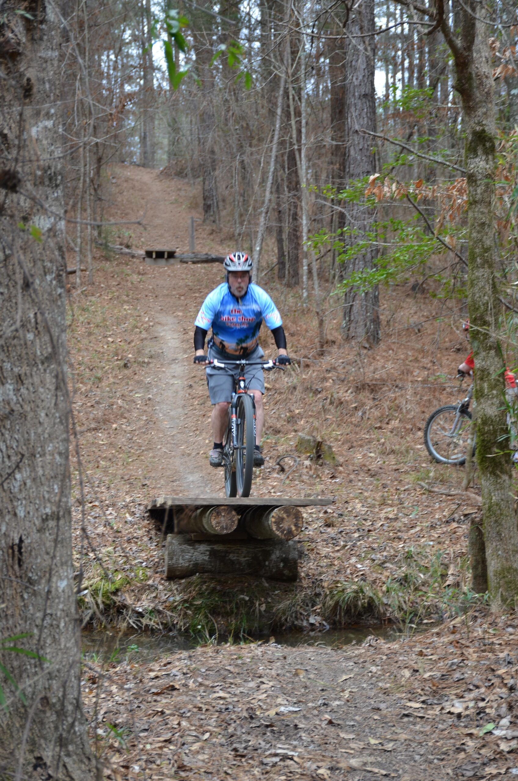 A mountain biker balancing on a narrow wooden bridge over a small creek, surrounded by a forested trail with trees and fallen leaves. Another bike is visible in the background on the path. Mt. Zion Bike Trails mountain bike trail.