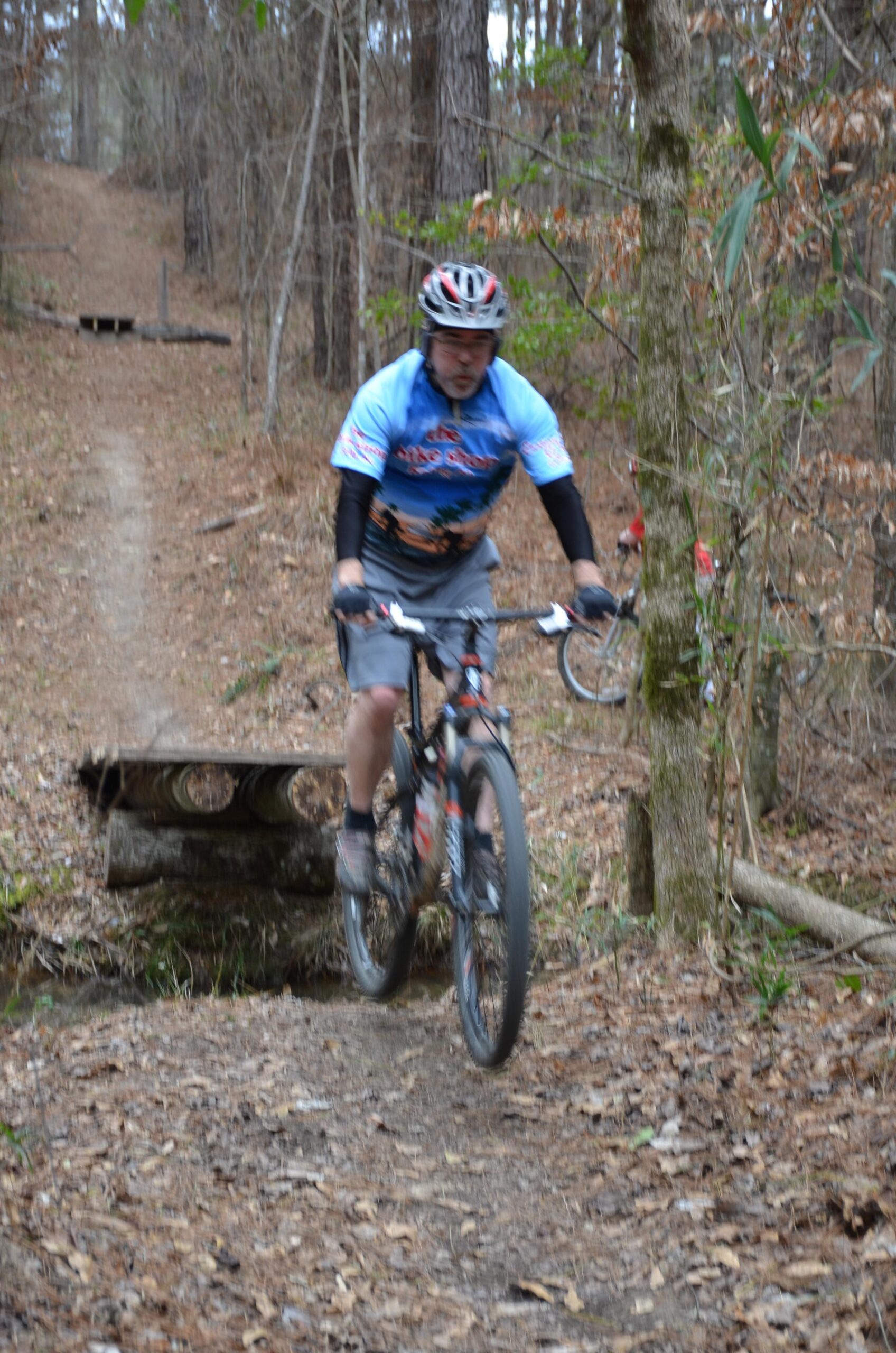 A mountain biker wearing a blue and black jersey and shorts rides over a small wooden bridge on a forest trail covered with fallen leaves. In the background, another cyclist can be seen in a red shirt. The scene is set in a wooded area with tall trees and a winding path. Mt. Zion Bike Trails mountain bike trail.