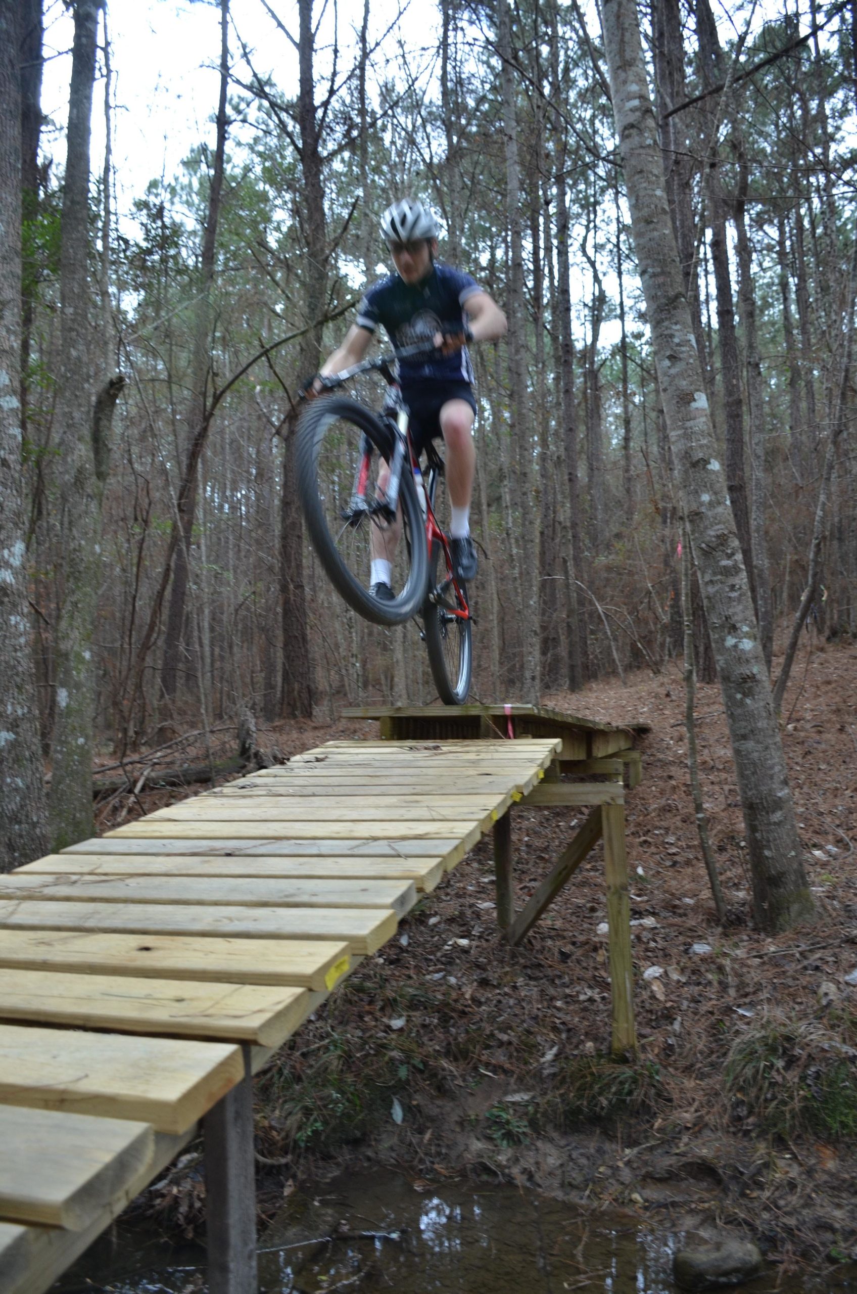 A mountain biker performing a jump off a wooden bridge in a forested area, showcasing the dynamic movement and thrill of the sport. Trees line the background, and the bike is captured mid-air above a shallow creek. Mt. Zion Bike Trails mountain bike trail.