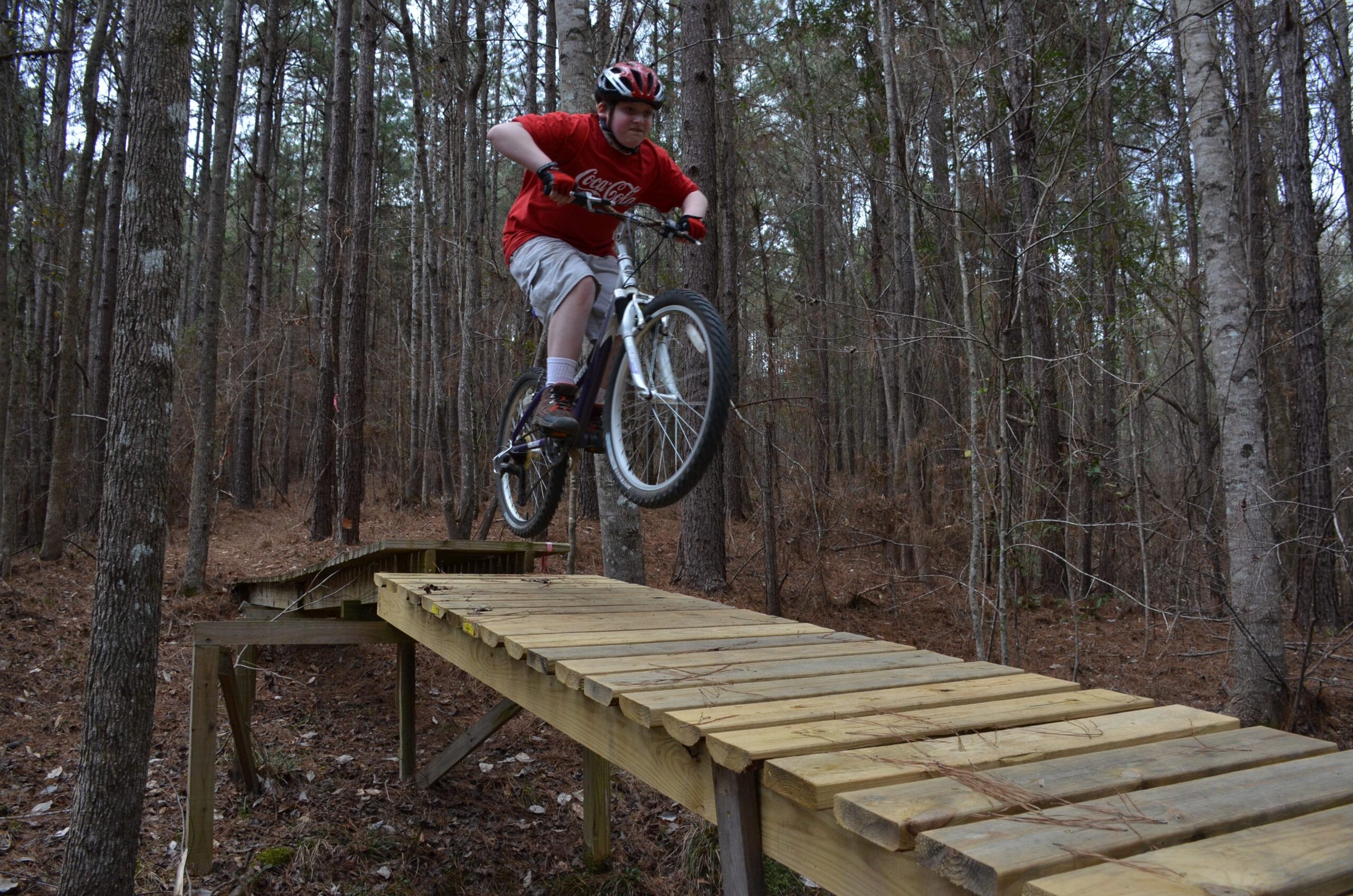 A young cyclist wearing a red t-shirt and a helmet is performing a jump on a mountain bike off a wooden plank bridge in a forested area. Surrounding trees create a natural backdrop as the rider catches air above the bridge. Mt. Zion Bike Trails mountain bike trail.