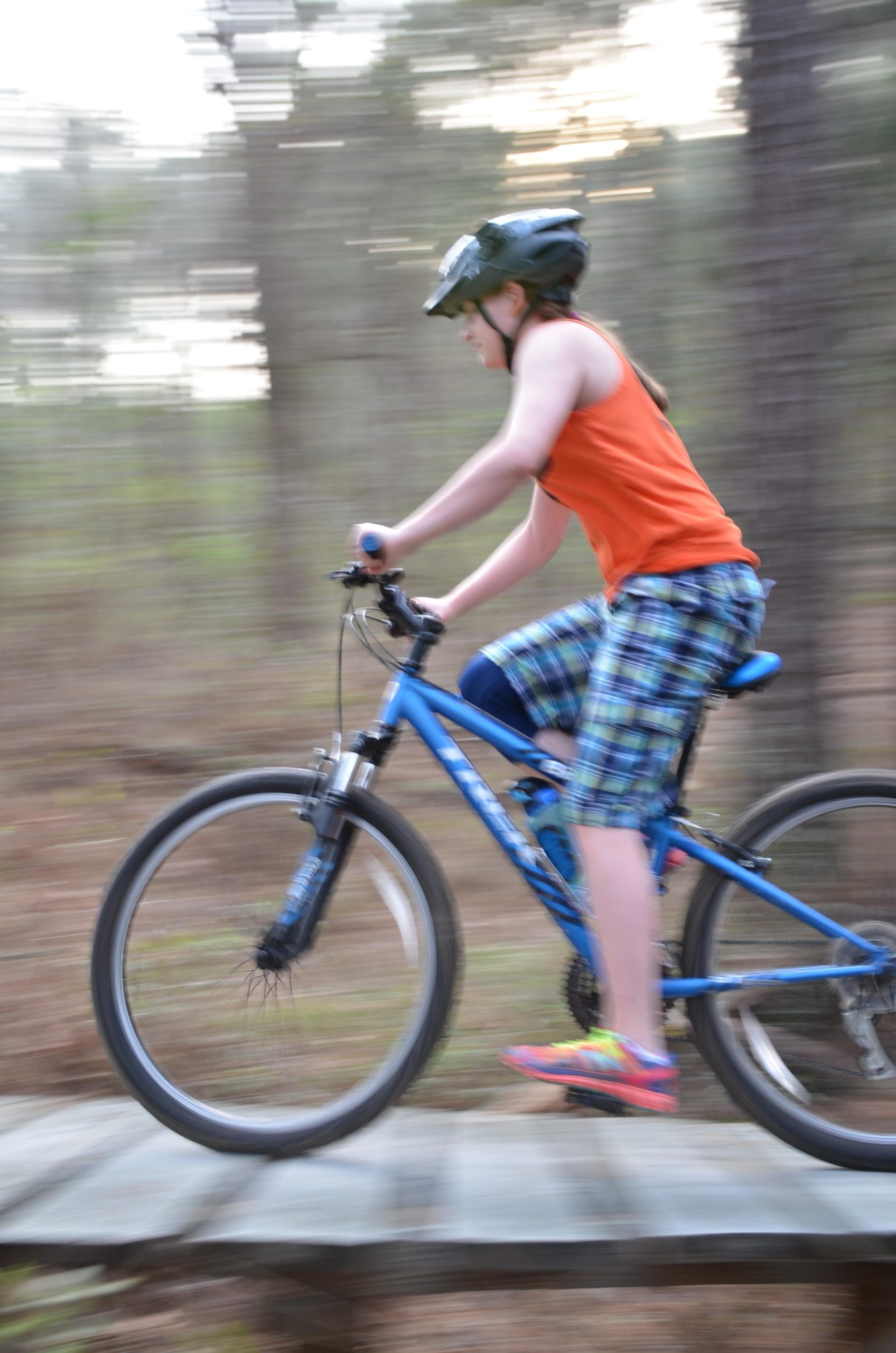 A child in an orange tank top and plaid shorts rides a blue mountain bike over a wooden bridge in a forested area, captured in motion with a blurred background to convey speed. The child is wearing a helmet for safety and colorful sneakers. Mt. Zion Bike Trails mountain bike trail.
