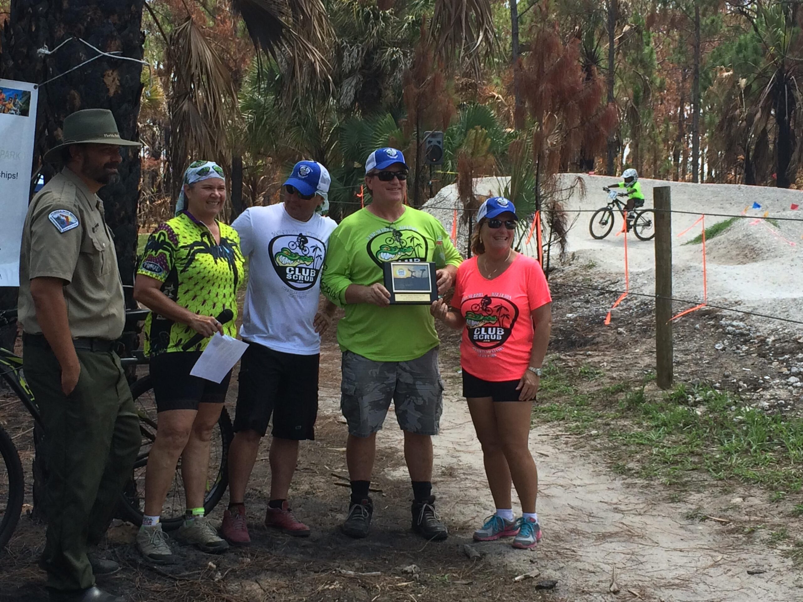 A group of five people stands together outdoors, celebrating an event related to mountain biking. Two of the individuals hold a plaque, and they are dressed in bright, themed shirts promoting "Club Scrub." A park ranger in uniform is present on the left. In the background, a child rides a bike on a dirt trail designed for cycling, surrounded by trees. Jonathan Dickinson State Park mountain bike trail.