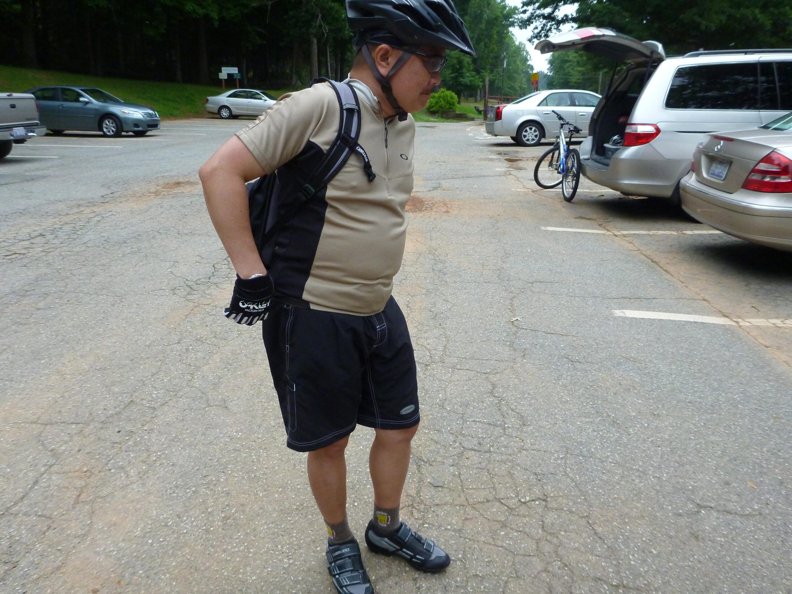 An individual wearing a cycling helmet, gloves, and athletic clothing, standing in a parking area with cars and a parked bicycle in the background. The person appears to be preparing for cycling, with a backpack and a focused demeanor. Country Park mountain bike trail.
