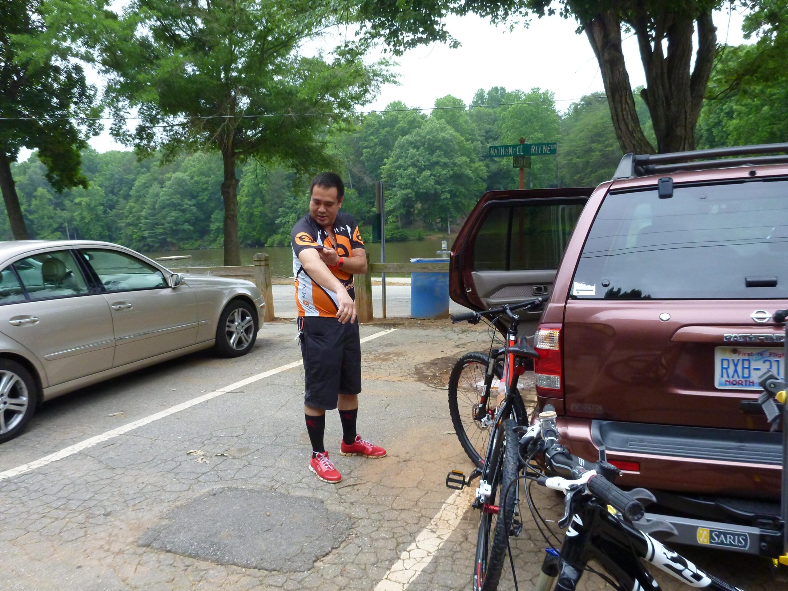 A person wearing a cycling jersey and shorts stands near a brown SUV parked by a lake, gesturing or adjusting their arm. Two bicycles are positioned next to the vehicle, and trees and greenery are visible in the background. A street sign indicating a nearby street can also be seen. Country Park mountain bike trail.