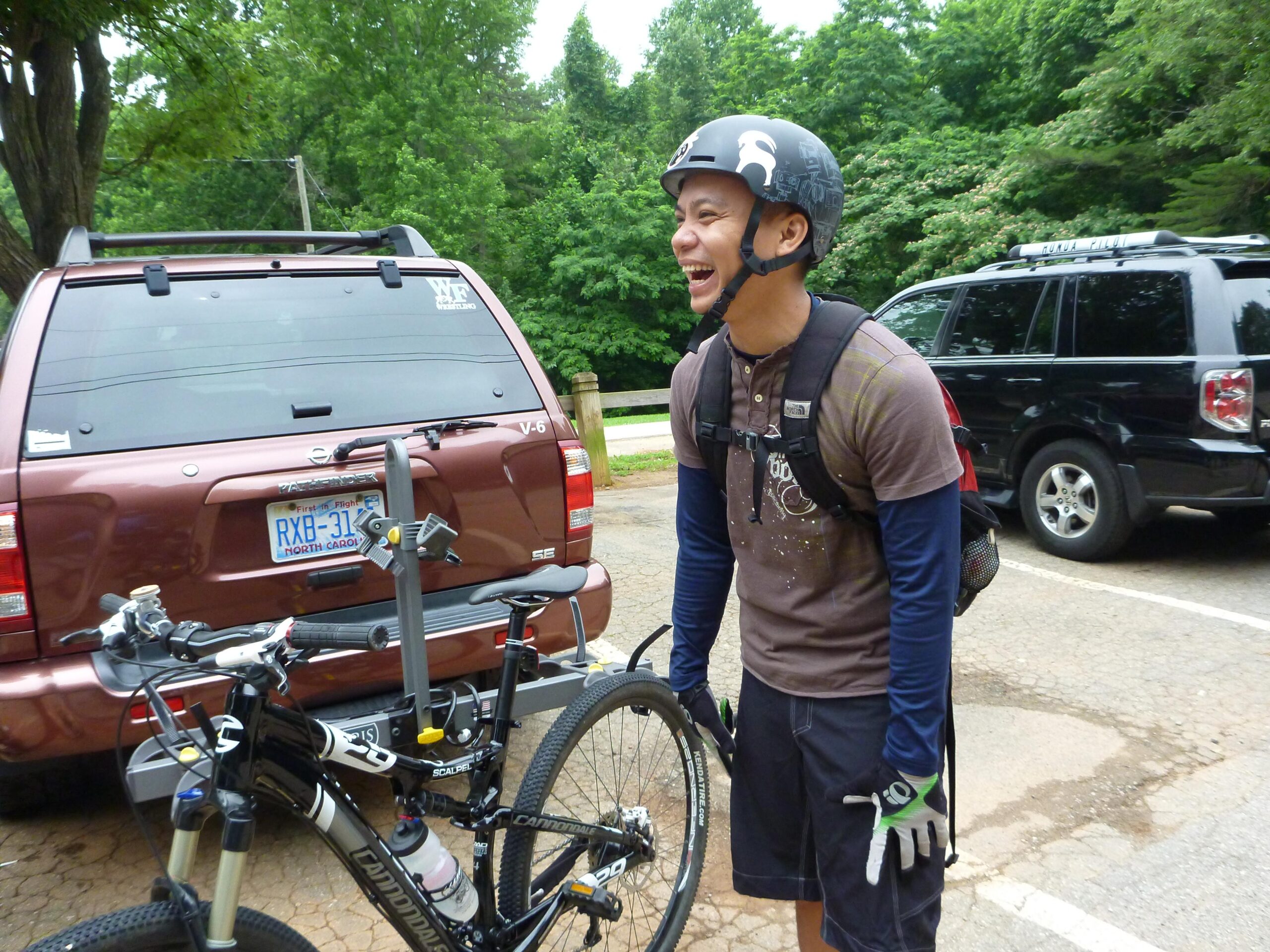 A smiling cyclist wearing a helmet and gloves stands next to a mountain bike in a parking area, surrounded by trees and vehicles. Country Park mountain bike trail.