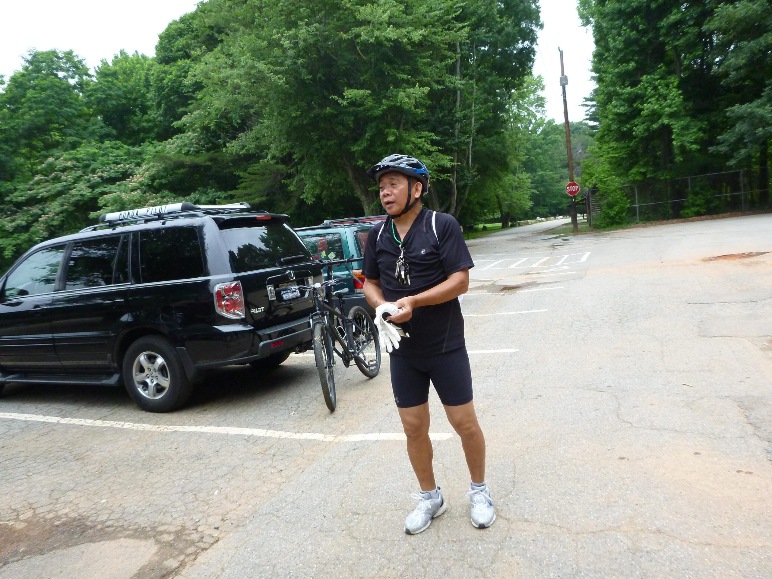 A cyclist in a black outfit and helmet stands beside a parked bicycle in a wooded area. They are holding gloves and appear to be preparing for a ride. Several cars are parked nearby, and a stop sign is visible in the background. The scene is set on a paved surface surrounded by greenery. Country Park mountain bike trail.
