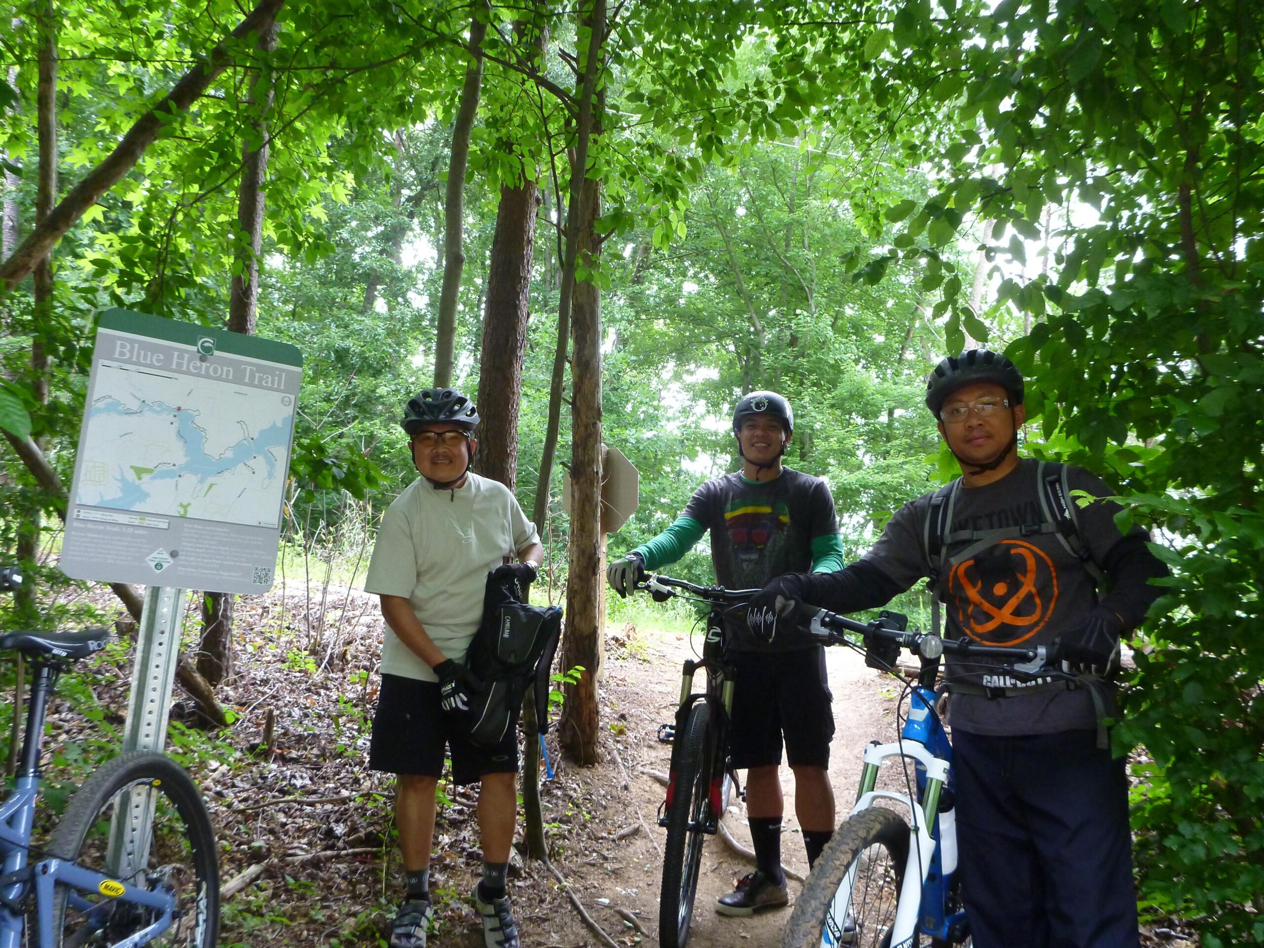 Three mountain bikers stand on a wooded trail next to a sign for the Blue Heron Trail. They are wearing helmets and cycling gear, and two of them are holding their bikes. The surroundings are lush with greenery, indicating a natural outdoor environment. Blue Heron mountain bike trail.