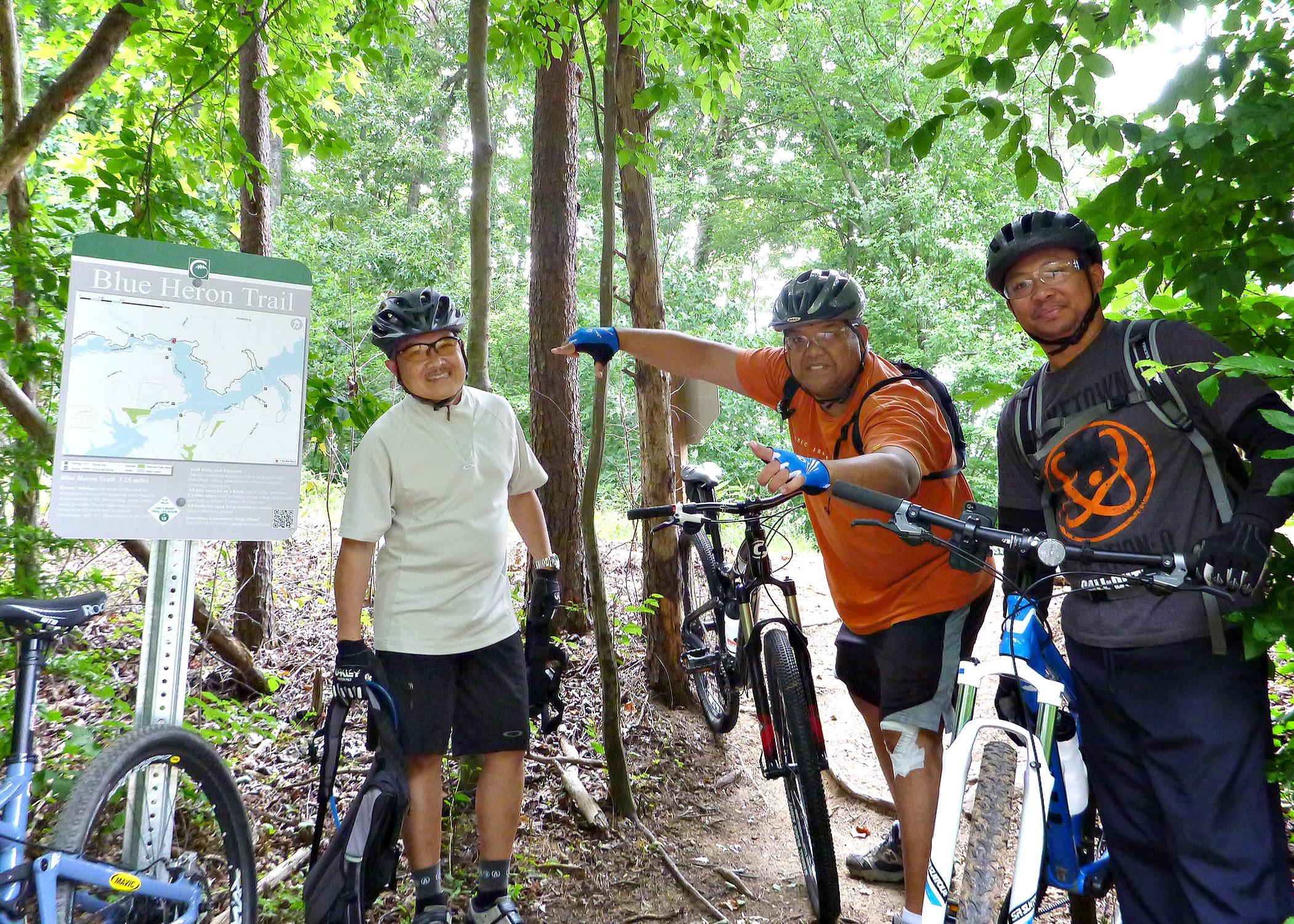 Three cyclists stand next to each other on a trail, posing for the camera. They are wearing helmets and casual biking attire, with two of them holding their bikes. A sign for the "Blue Heron Trail" is visible beside them, displaying a map of the area. The background features lush green trees and foliage. Blue Heron mountain bike trail.