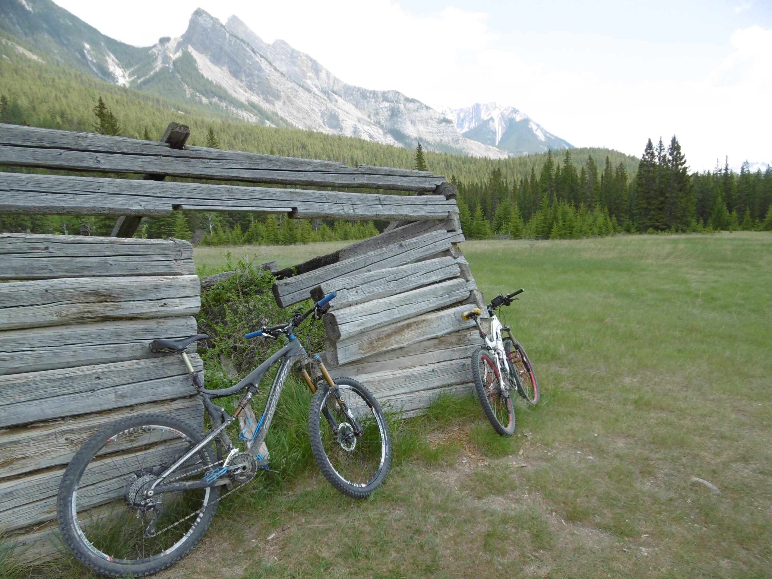 Two mountain bikes, one gray and one white, are leaning against a weathered wooden structure in a grassy area. In the background, there are tall green trees and a mountain range under a partly cloudy sky. The scene conveys a sense of outdoor adventure and exploration. The Overlander mountain bike trail.