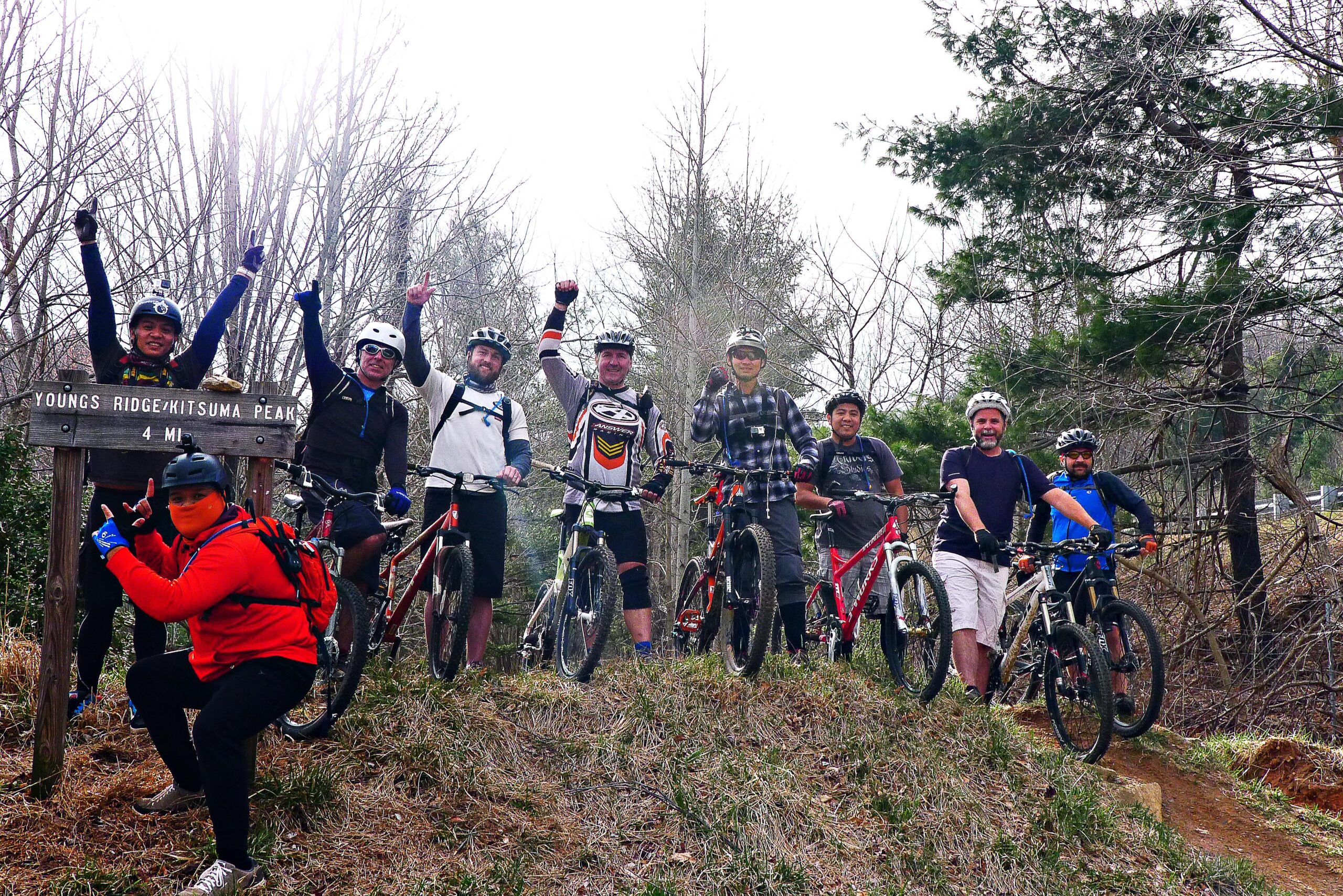 Group of eight mountain bikers celebrating at a trail sign for Youngs Ridge/Kitsuma Peak, with a distance of 4 miles displayed. They are posing on a grassy incline, wearing helmets and cycling gear, with some individuals making peace signs and cheering. The background features bare trees and hints of greenery, indicating an outdoor setting. Kitsuma mountain bike trail.