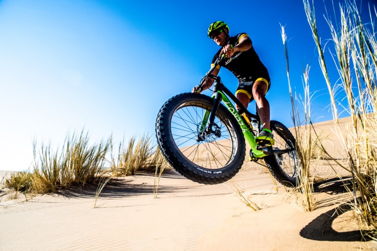 A cyclist performing a jump on a fat bike in a sandy desert landscape, with blue skies and tall grass in the foreground. The rider is wearing a helmet and athletic gear, showcasing an action-packed scene.