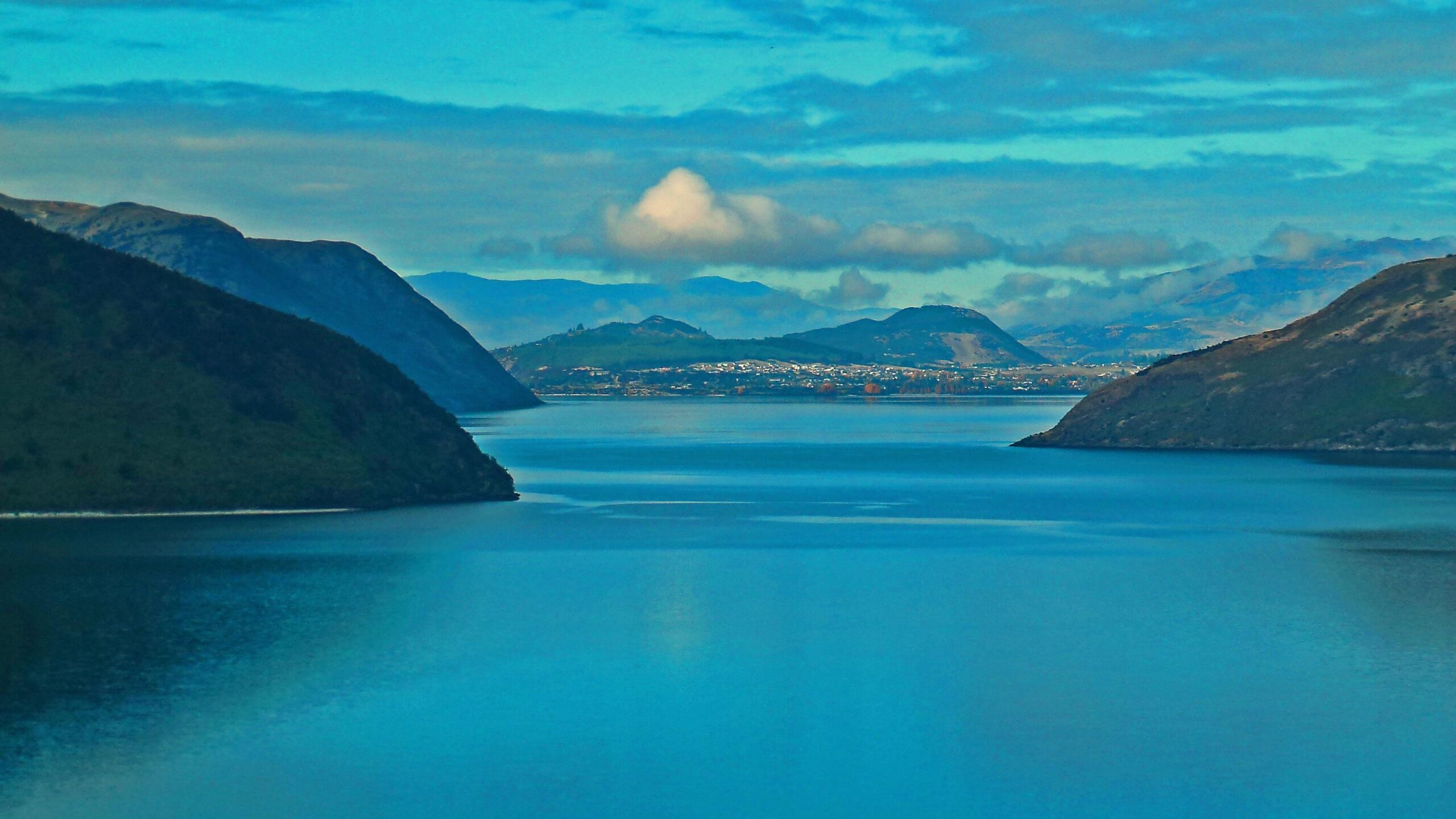 A serene view of a tranquil body of water surrounded by hills and mountains under a partly cloudy sky. The water reflects the blue tones of the sky, while distant land is visible, featuring trees and buildings along the shoreline. Minaret Burn Track mountain bike trail.