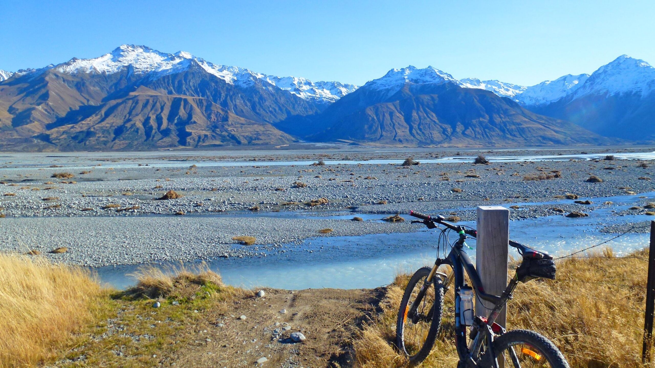 A mountain bike resting against a post along a gravel trail, with a river flowing nearby and snow-capped mountains in the background under a clear blue sky. Tasman River Track mountain bike trail.