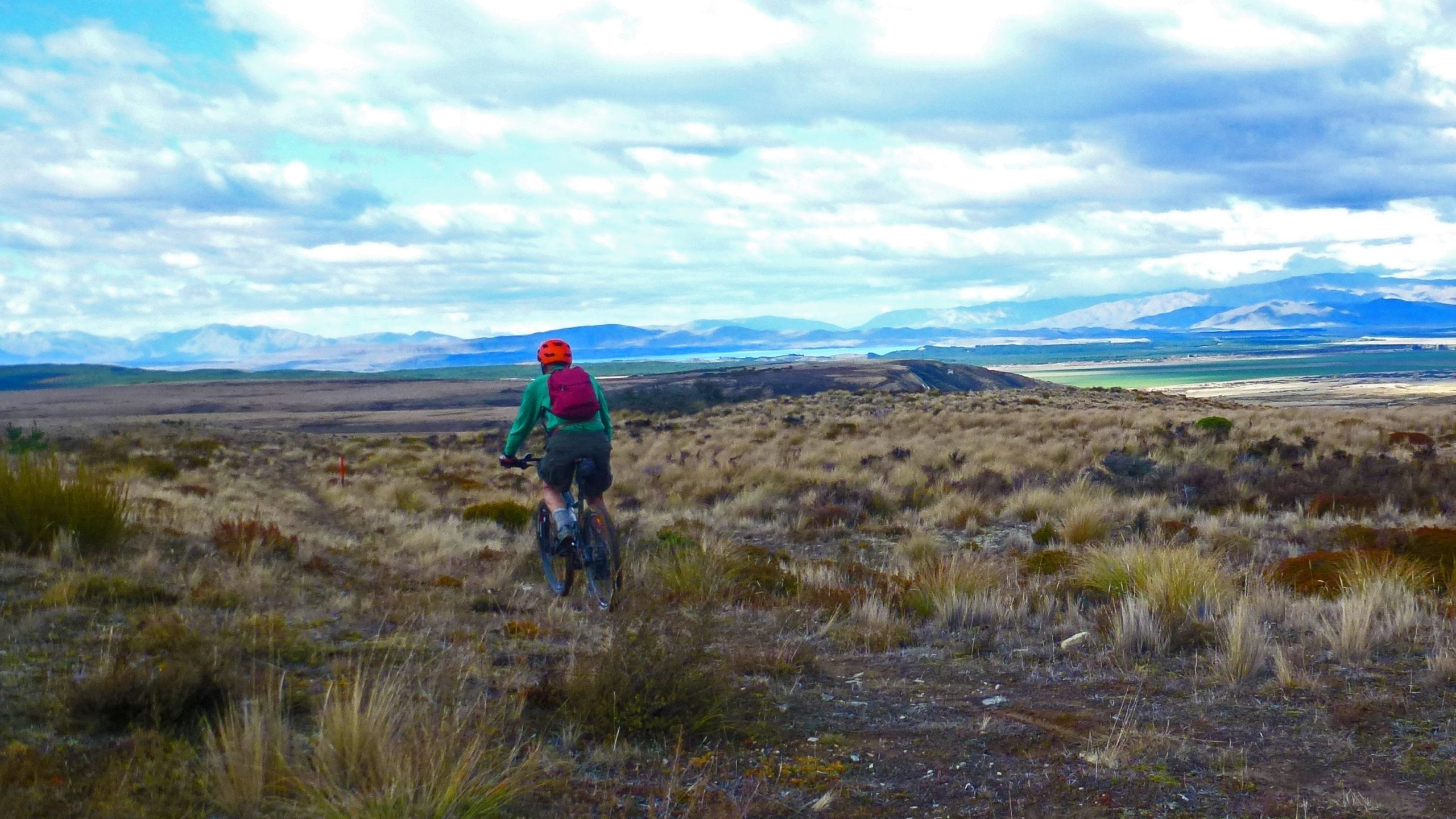 A person riding a mountain bike along a rugged trail, surrounded by golden grasses and shrubs, with distant mountains and a scenic landscape in the background under a partly cloudy sky. Dusky Trail mountain bike trail.