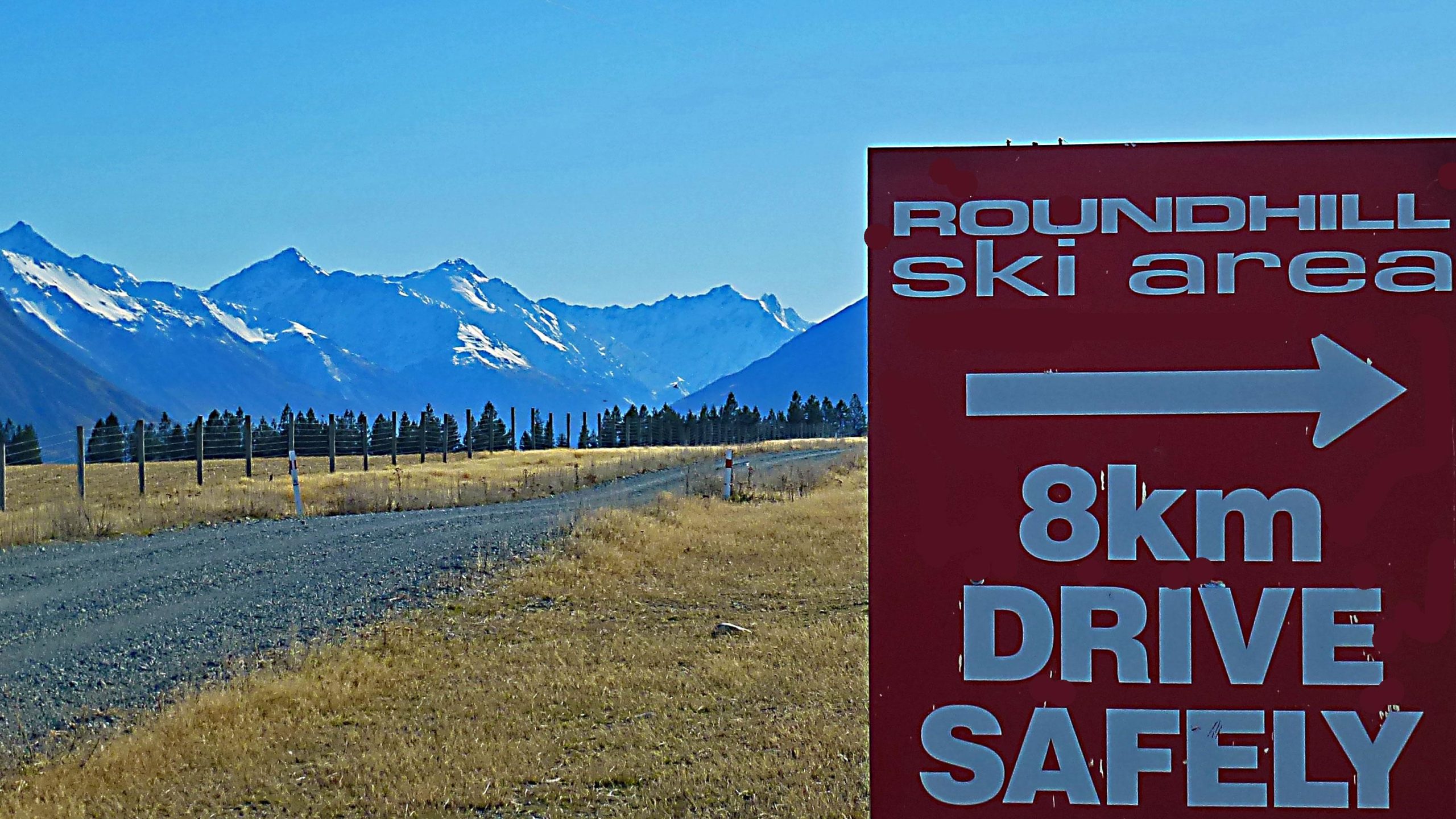 A red sign indicating the direction and distance to Roundhill Ski Area, with the text "8km DRIVE SAFELY" prominently displayed. In the background, there are snow-capped mountains under a clear blue sky, with a dirt road leading towards the sign and surrounding grassy fields. Richmond Trail Loop mountain bike trail.