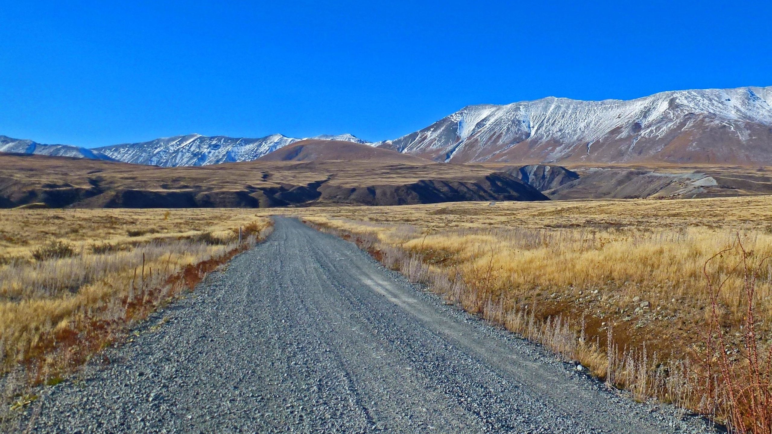 A gravel road stretches through a wide, open landscape surrounded by brown hills and snow-capped mountains under a clear blue sky. The foreground features dry grasses and sparse vegetation, leading the viewer's eye towards the distant mountain range. Richmond Trail Loop mountain bike trail.