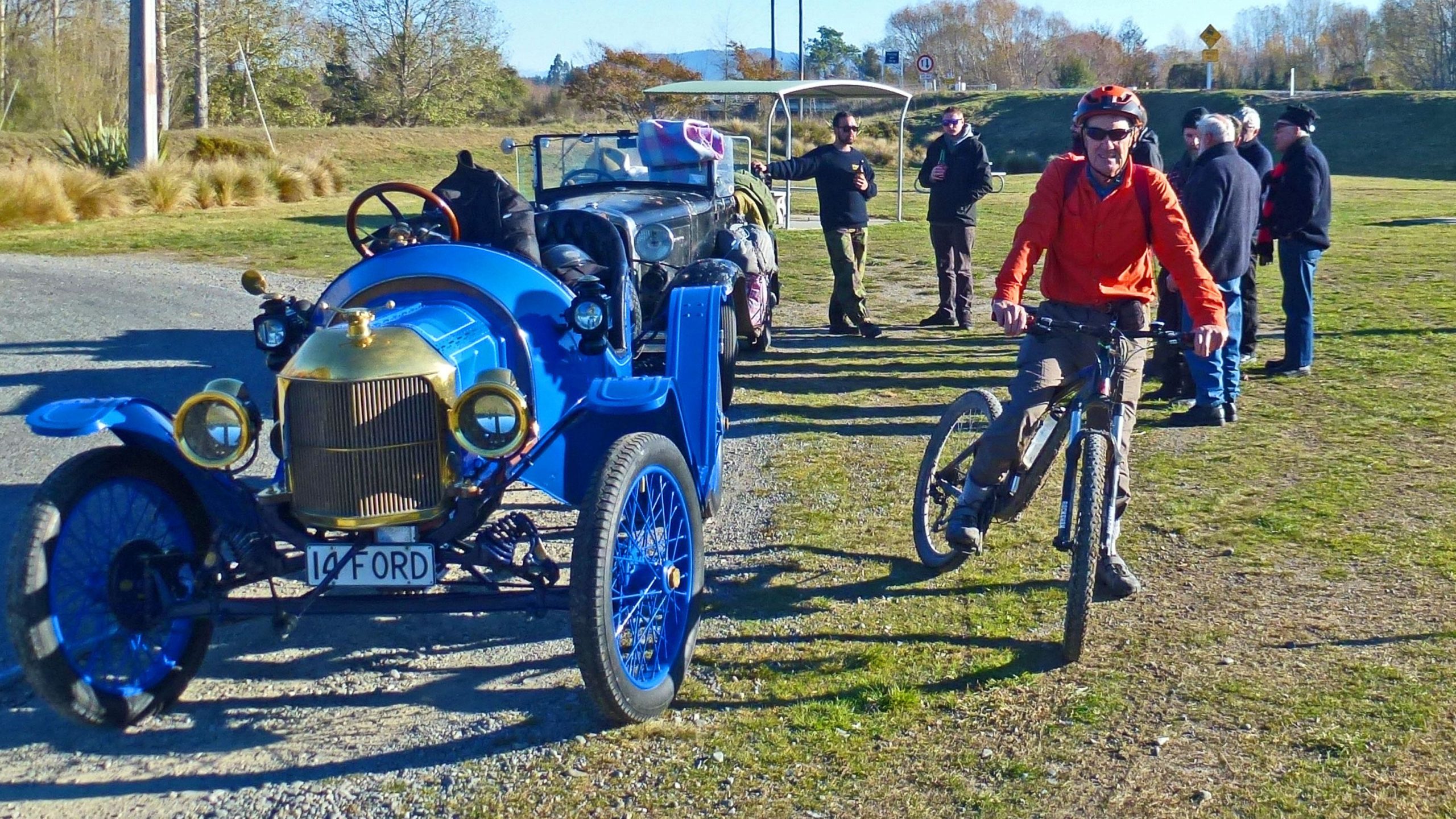 A vintage blue car with a shiny gold grille is parked on a grassy area, while a man in an orange jacket rides a mountain bike nearby. A small group of people stands in the background, chatting and enjoying drinks, with trees and a clear sky visible in the surroundings. Geraldine MTB Track mountain bike trail.