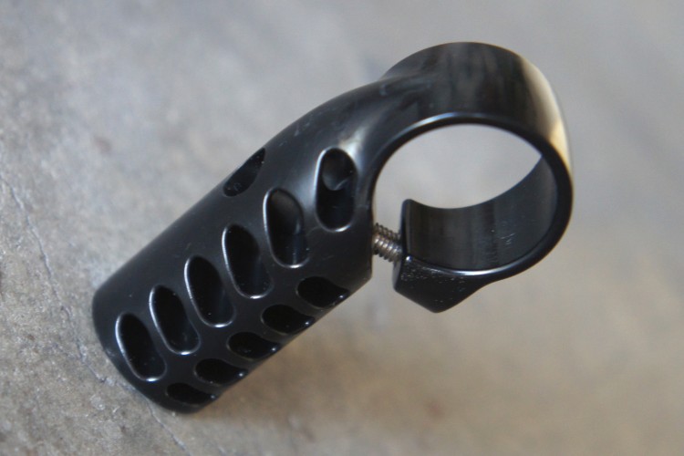 A close-up of a black plastic bike bell or handlebar accessory featuring a hollow, curved design with multiple ventilation holes. The item is sitting on a textured gray surface, showcasing its glossy finish and detailing.