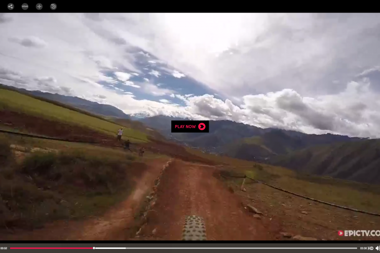 A mountain biking scene showing a dirt trail winding through a picturesque landscape of hills and valleys under a partly cloudy sky. The image captures the view from the biker's perspective, with the bike tire in the foreground and other riders in the distance. A play button is overlaid, indicating that it is a video still.
