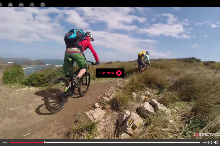 Two mountain bikers riding along a scenic dirt trail surrounded by grass and rocks, with a coastal view in the background and a blue sky dotted with clouds. A play button overlay is present in the center of the image.