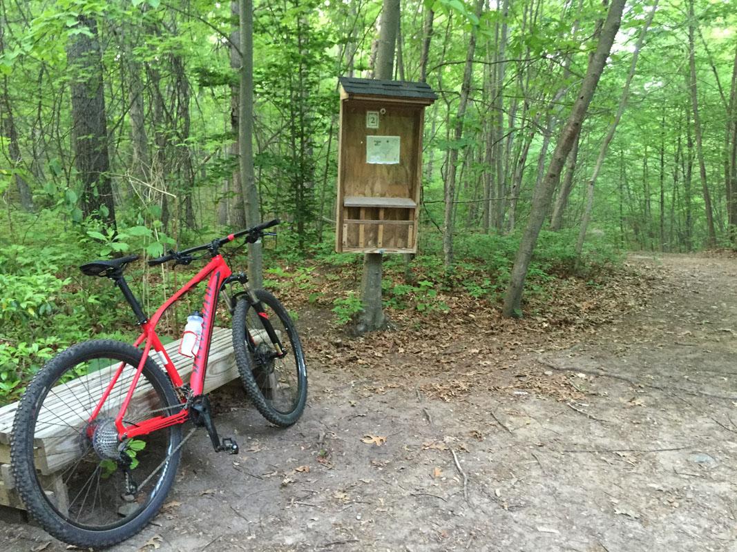 A red mountain bike parked on a wooden bench beside a trail in a green forest. A wooden information kiosk is visible in the background, surrounded by trees and foliage. The ground is covered with dirt and fallen leaves. Rockland Preserve mountain bike trail.