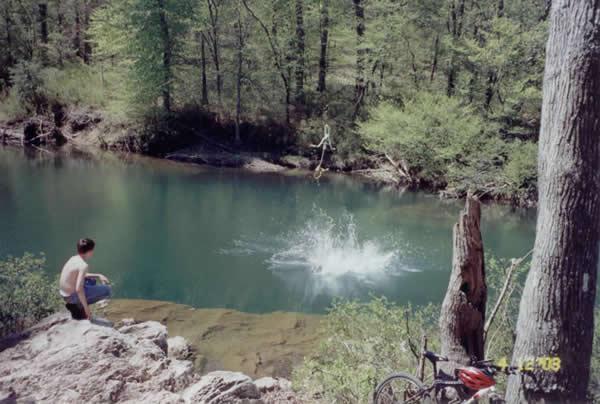 A scenic view of a natural swimming hole surrounded by trees. In the foreground, a person in swim trunks stands on a rocky edge, observing. In the background, another person is mid-air above the water, about to make a splash. A bicycle is parked nearby on the right side of the image. The vibrant green foliage reflects the warm, sunny atmosphere. Brush Heap Loop mountain bike trail.